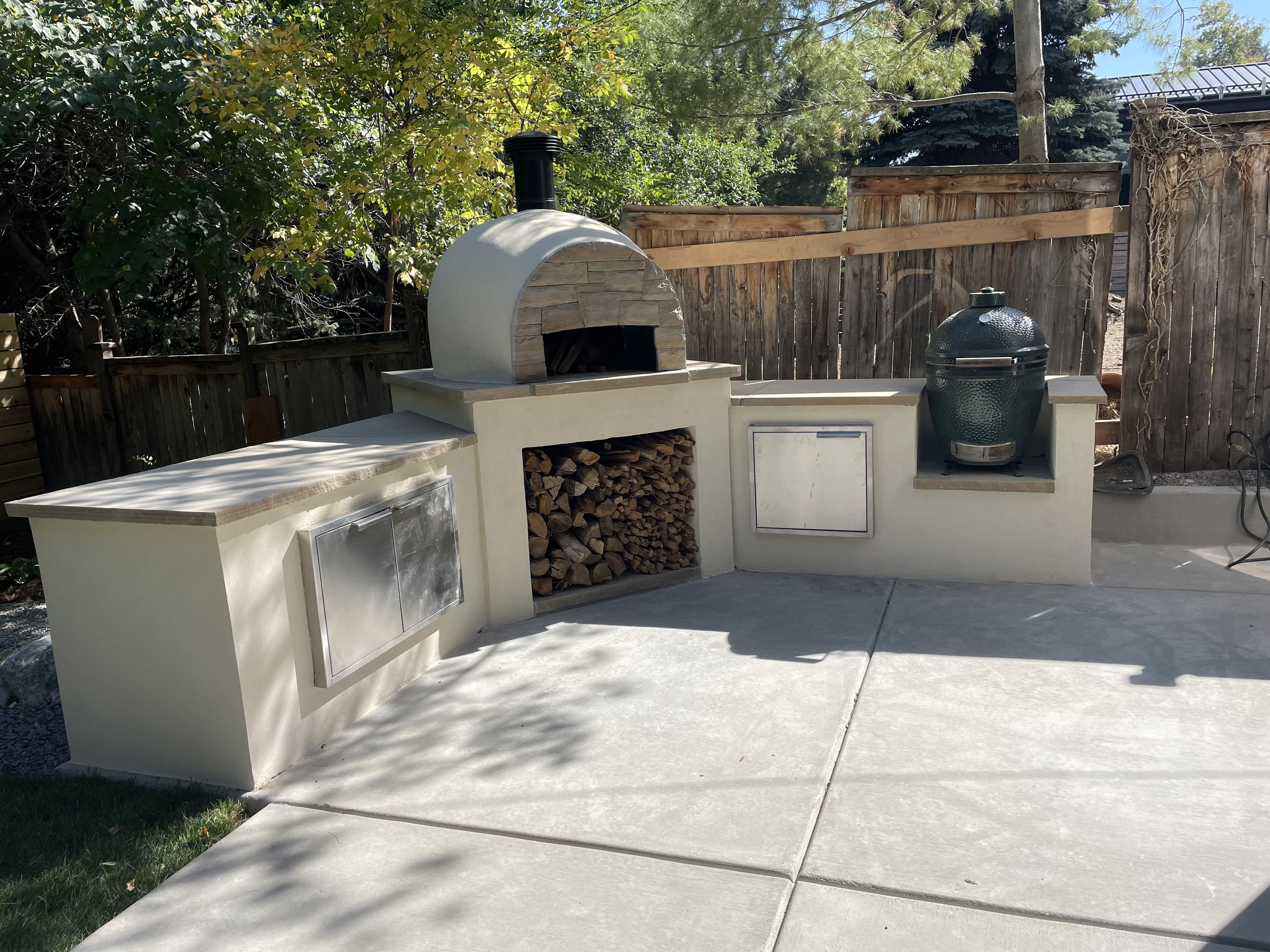 Custom outdoor stone kitchen with wood-fired oven, wood storage, and a decorated grill in a backyard.
