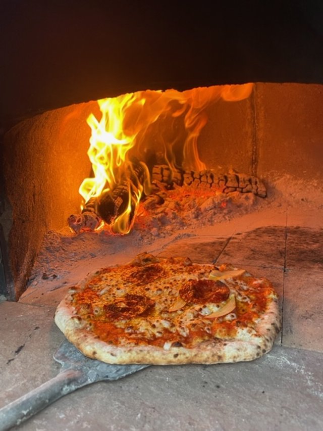 A pizza being baked in a custom built stone wood-fired oven with flames visible in the background.