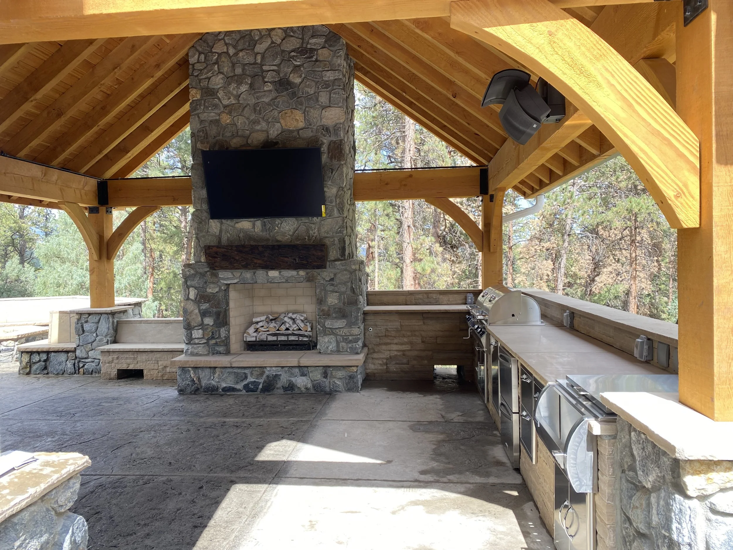 Custom outdoor stone fireplace with a mounted flat-screen TV above, open kitchen area with grill and counter space, surrounded by wooden beams and trees in the background crafted by Boulder StoneScapes.