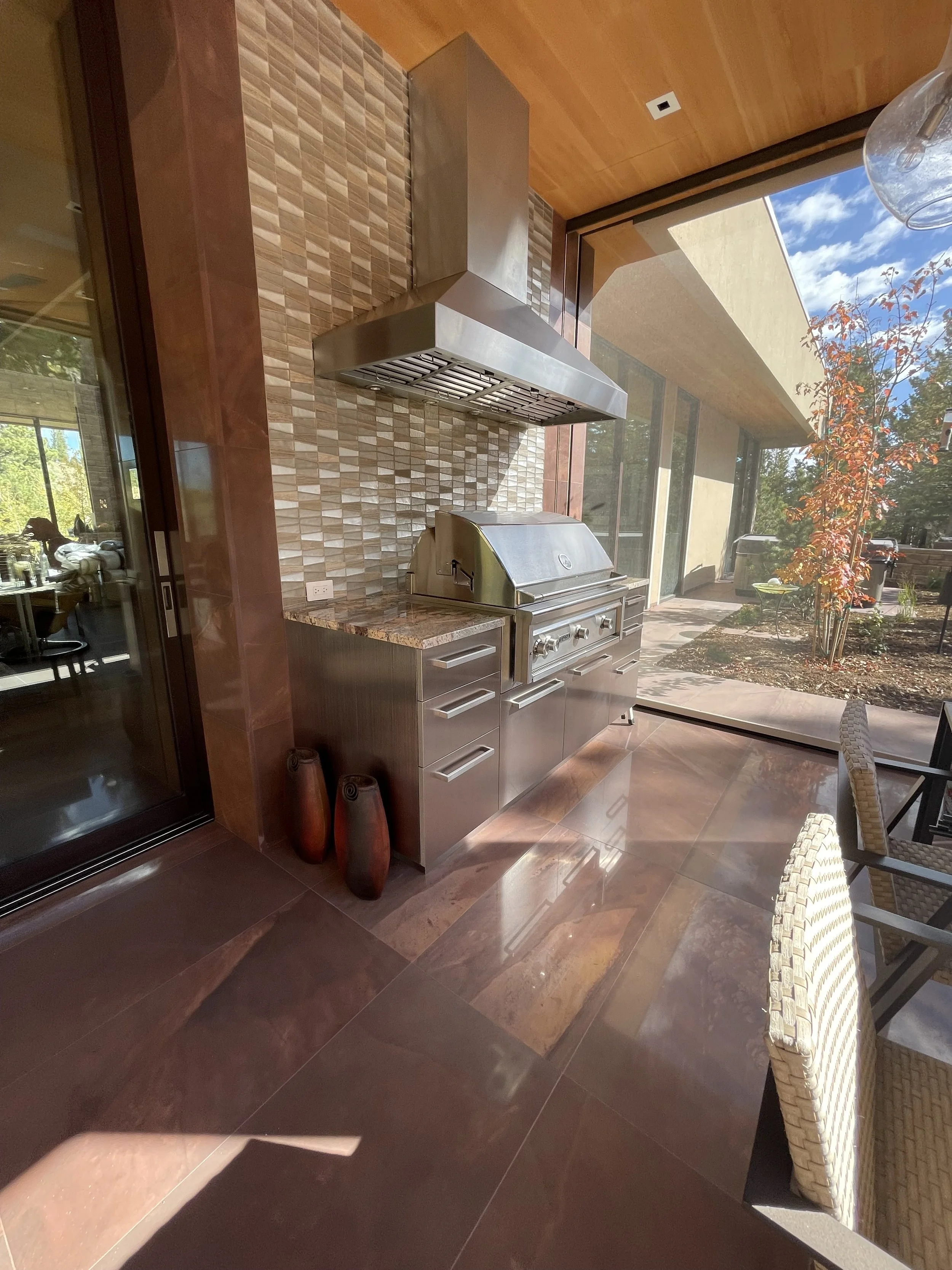 Outdoor kitchen with stainless steel grill, chimney, countertop, and decorative vases, beside large glass sliding door and patio furniture, with garden and trees outside.