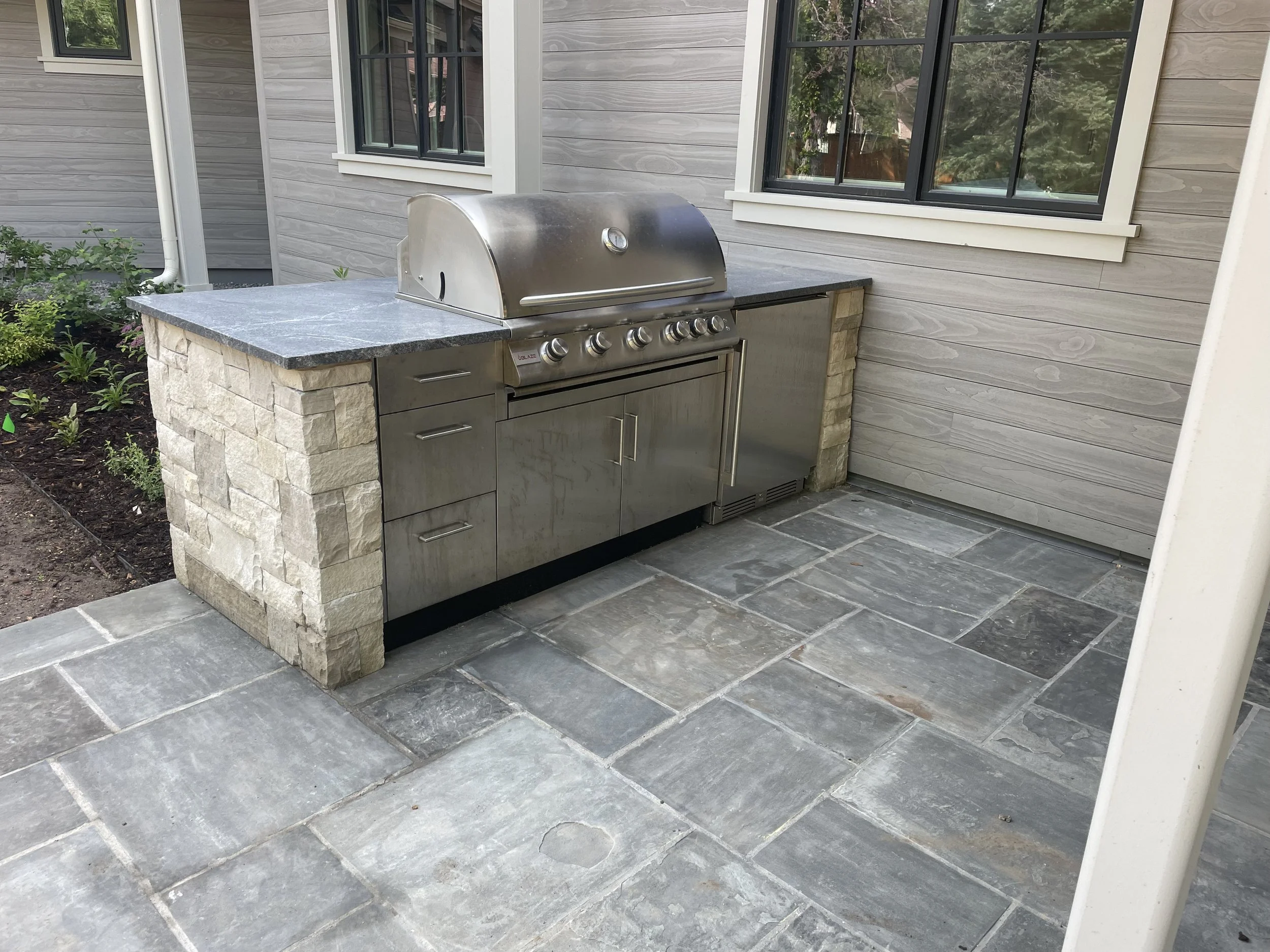 Outdoor stone patio area with a stainless steel gas grill and countertop, adjacent to a house with gray wooden siding and two black framed windows.