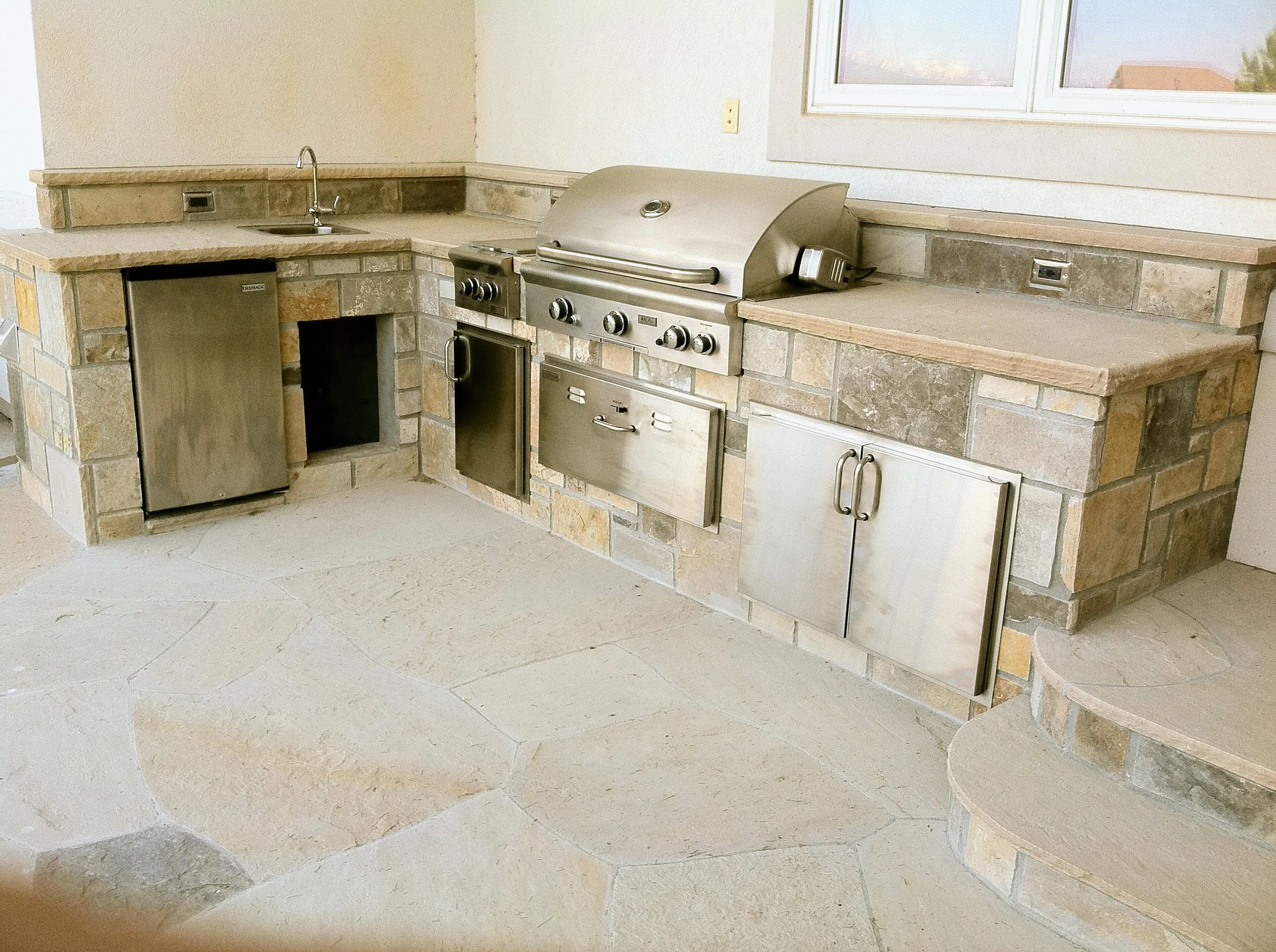 Outdoor kitchen with custom brick and stone countertops, built-in stainless steel grill, mini fridge, and small sink, with large window in the background.