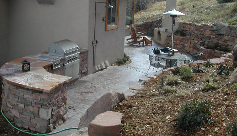 Outdoor patio area with a grill, table, chairs, an umbrella, and a chair, next to a house with a small window, surrounded by dirt and plants.