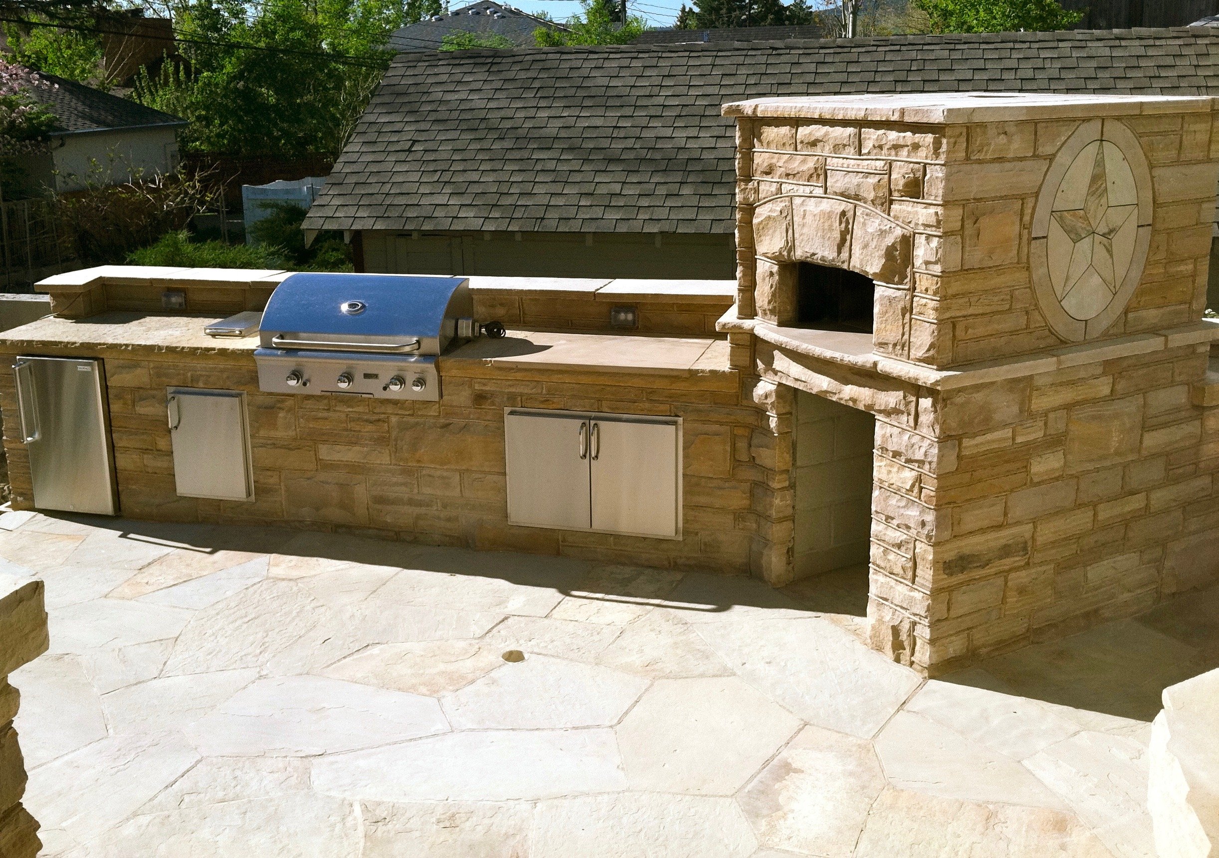 Outdoor built-in custom masonry stone grill and fireplace with metal storage cabinets, set on a stone patio.