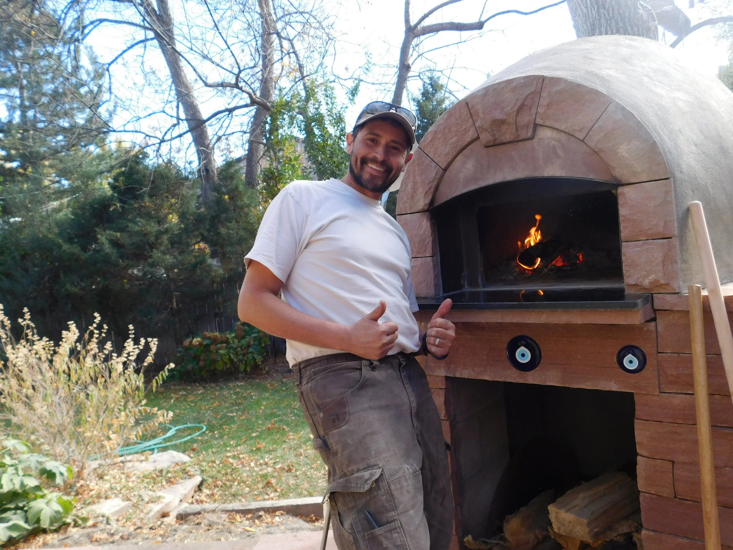 Brian Scott, owner of Boulder StoneScapes, smiling and giving a thumbs-up next to an outdoor pizza oven with a fire burning inside. He is wearing a white t-shirt, cargo shorts, sunglasses on his head, and a wedding ring.