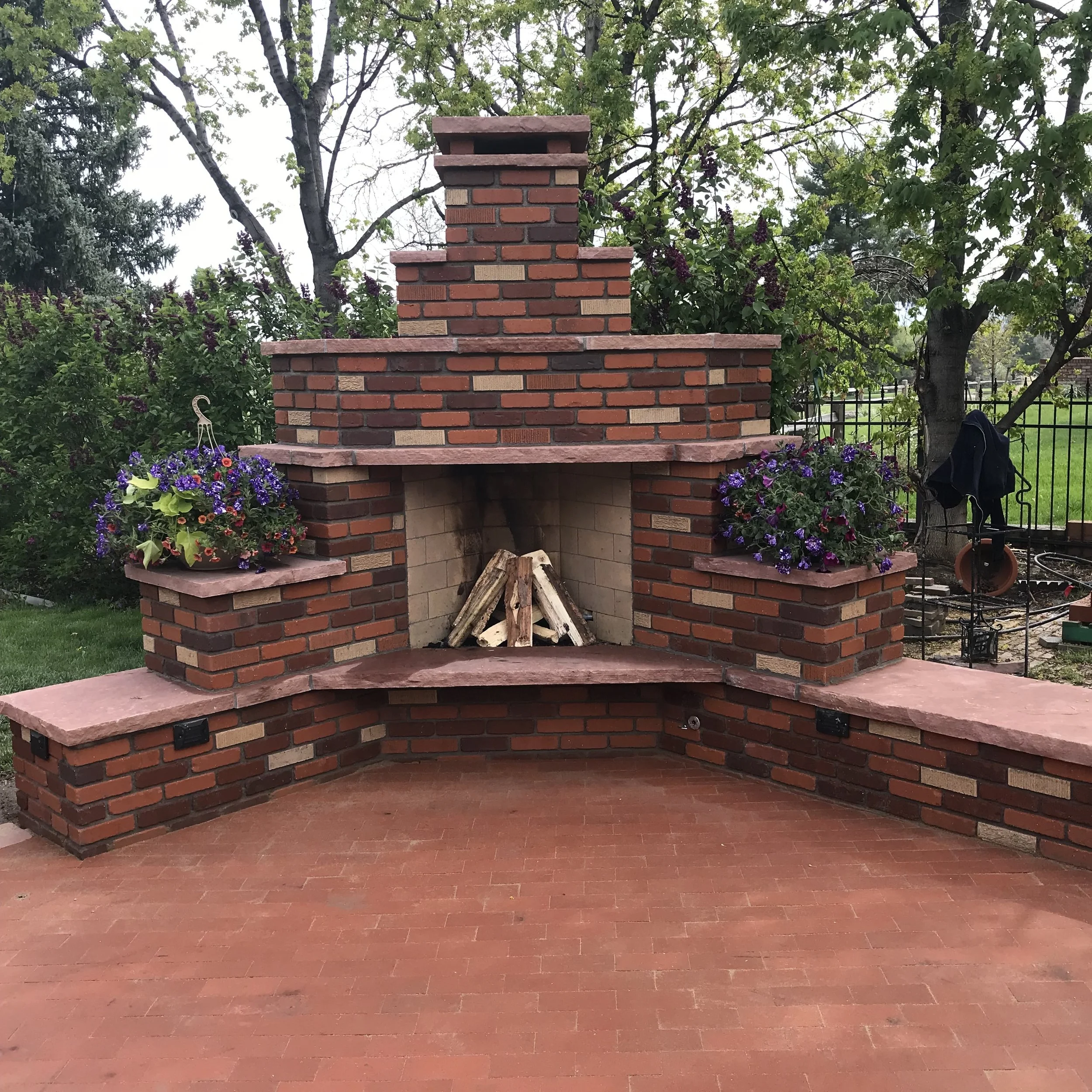 A custom stone outdoor fireplace with firewood stacked inside, flanked by two flower pots with purple and red flowers, set in a backyard with trees and a black iron fence.