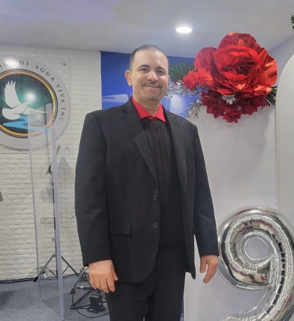 A man in a black suit with a red shirt and black sweater stands indoors in front of a decorative background featuring a large red paper flower and a silver balloon shaped as the number nine. There is a logo on the wall with a white dove and the text 'Protección de Agua Viva Inc.'