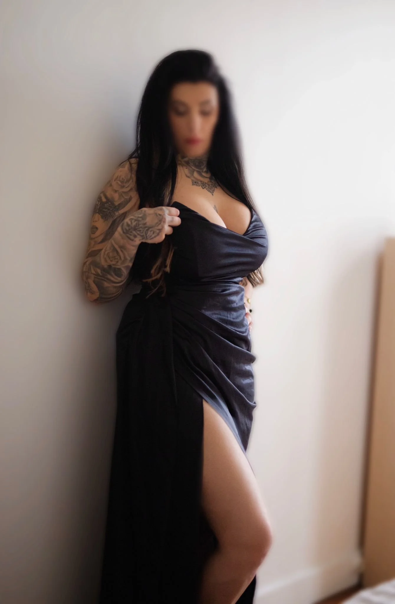 Femme avec tatouages portant une robe en satin noir, posant contre un mur, avec une robe dépassant par une fente.