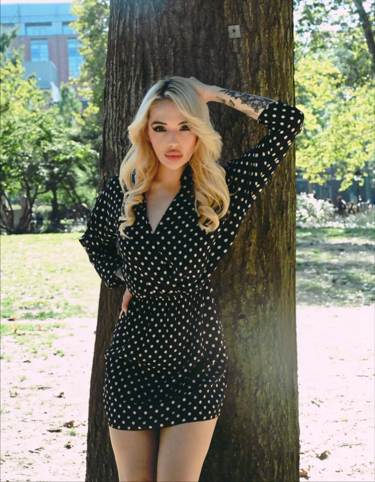 A woman with long blonde hair stands outdoors in a park, leaning against a tree. She is wearing a black dress with white polka dots and has her left hand behind her head. The background features lush green trees, bushes, and a cityscape with building