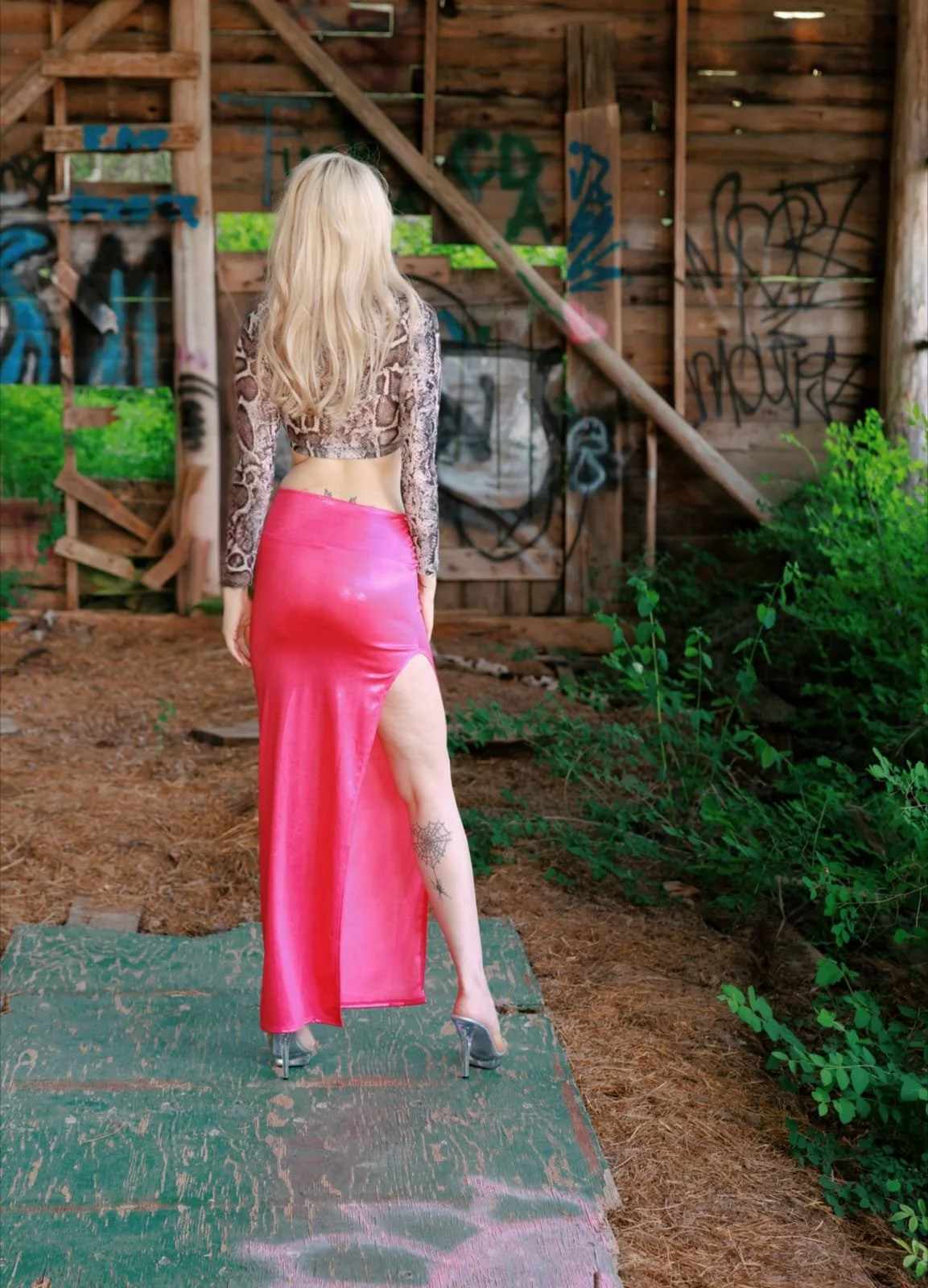 A woman with long blonde hair wearing a leopard print top, a pink satin skirt with a high slit, and silver high heels standing on a weathered green platform in front of a graffiti-covered wooden structure in an outdoor, overgrown area.