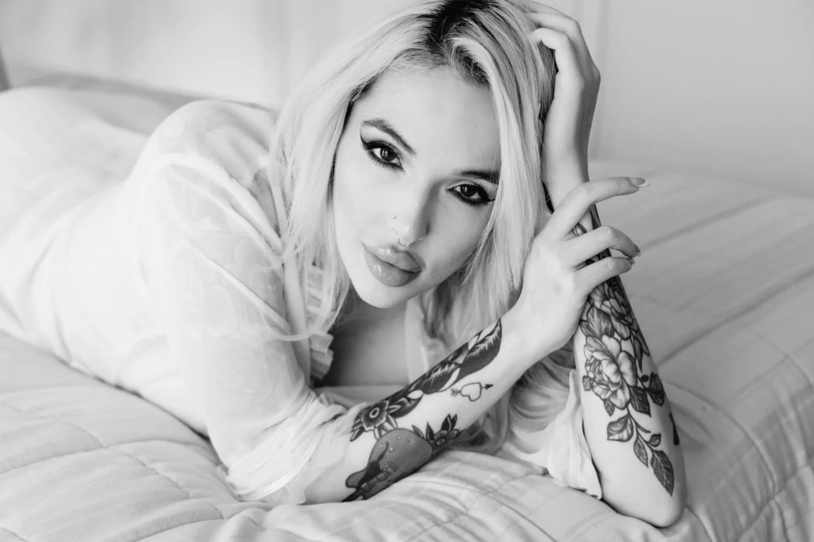 Black and white photo of a woman with blonde hair, tattoos on her arms, and a nose piercing, lying on a bed and looking at the camera.