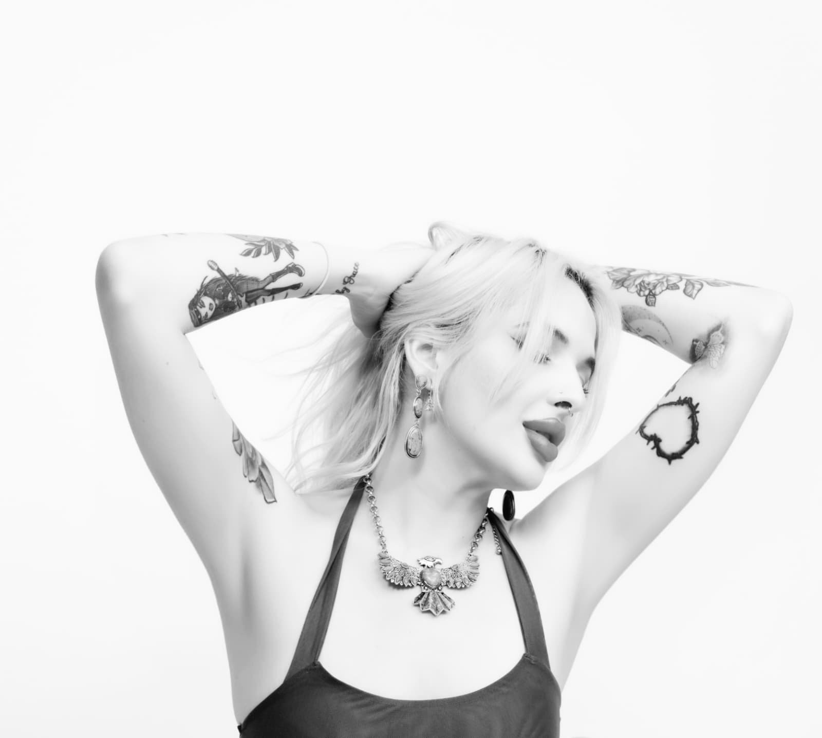 A young woman with light hair and tattoos on her arms, wearing earrings and a necklace, with her eyes closed and hands behind her head against a plain white background.