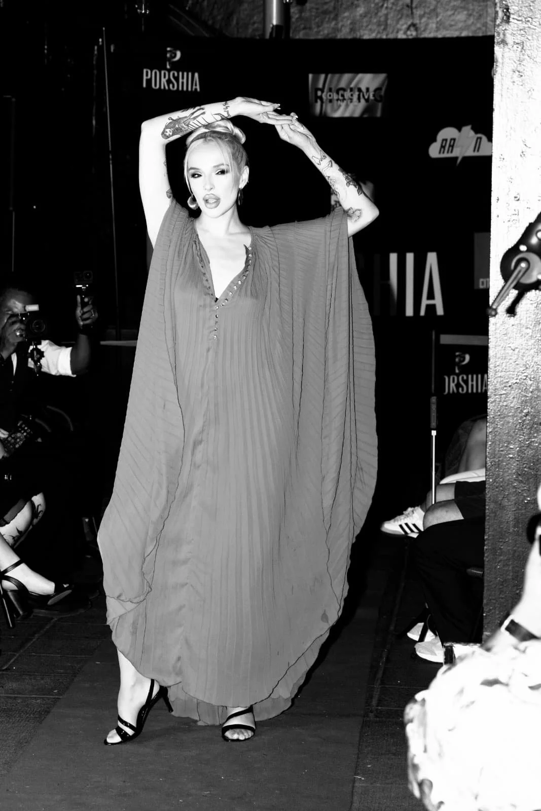 A woman walking on a runway during a fashion show, wearing a flowing pleated dress and high heels, with tattoos on her arms and earrings, while photographers and audience observe.