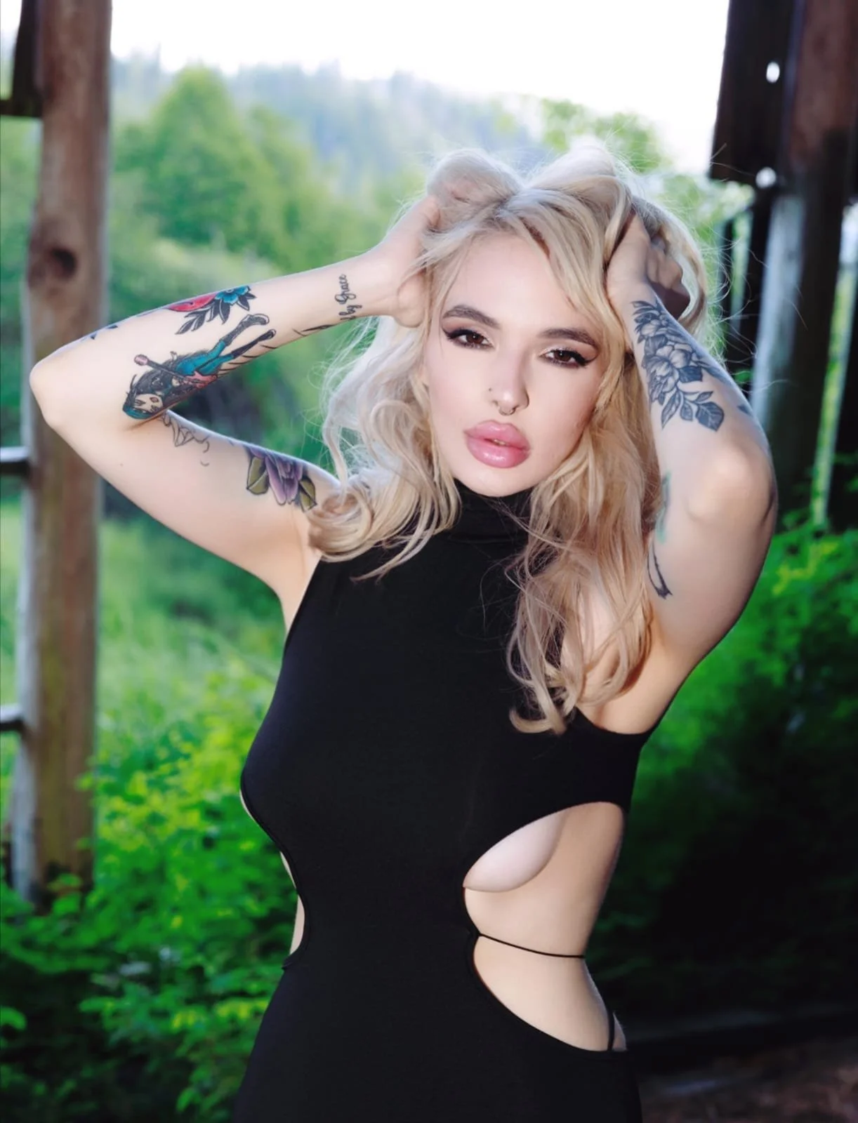 A woman with tattoos on her arms and long blonde hair standing outdoors near wooden posts with a blurred green forest background. She is wearing a black dress with cutouts.