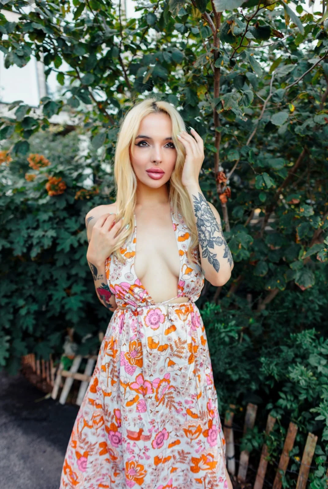 A blonde woman with tattoos on her arms, wearing a floral dress with a plunging neckline, standing outdoors in front of green leafy bushes.