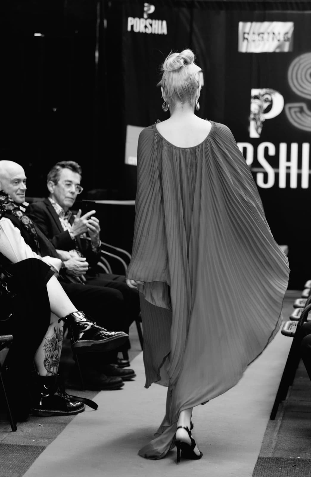 A woman with short blonde hair wearing a flowing, pleated gown and high-heeled shoes walking on a runway at a fashion show with seated audience members watching.