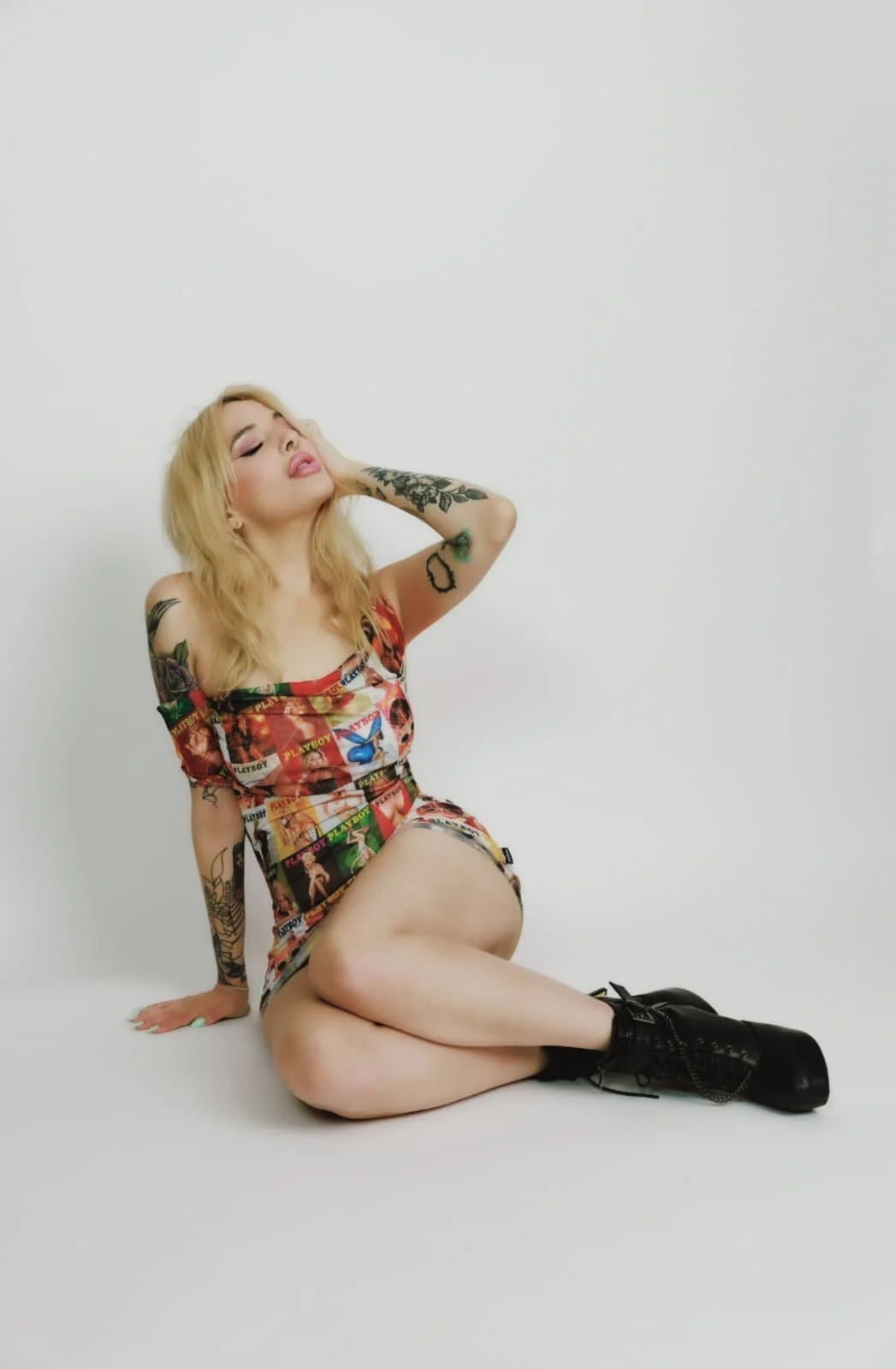 A woman with blonde hair and tattoos wearing a colorful, magazine collage-style dress, black boots, sitting on the floor with her legs crossed, eyes closed, and one hand touching her head against a plain white background.