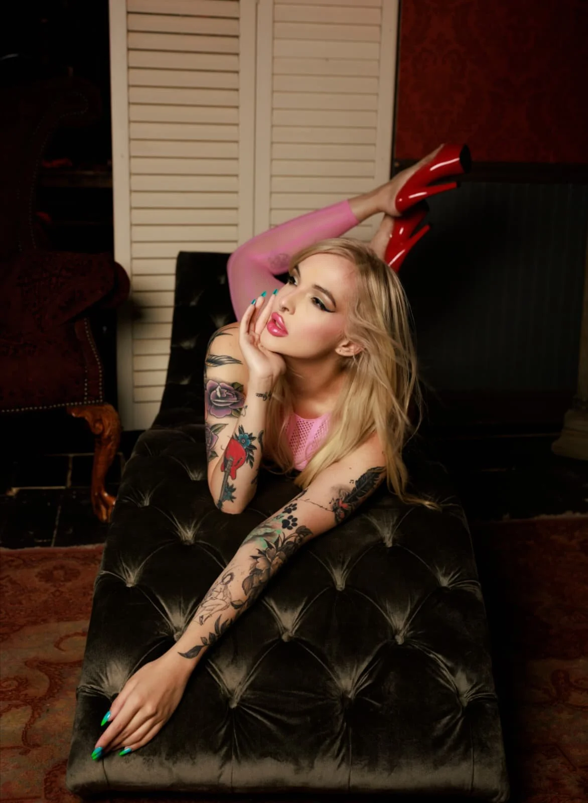 A woman lying on a black tufted ottoman, propped up on her elbows, wearing pink fishnet stockings and red high heels, with long blonde hair and colorful tattoos on her arms. She is resting her chin on her hand and gazing to the side.