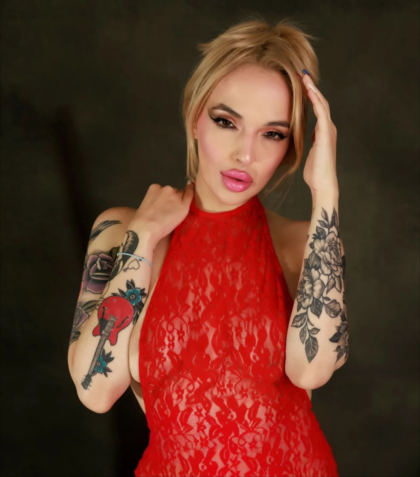 A woman with dyed blonde hair, tattoos on both arms, wearing a red lace dress, and posing against a dark background.