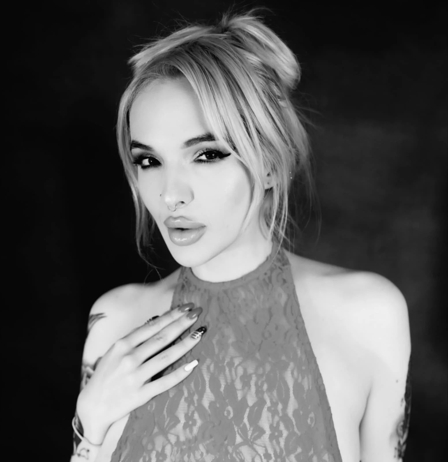 Black and white portrait of a woman with long, wavy hair, wearing a lace top, with her hand on her chest and a confident expression.