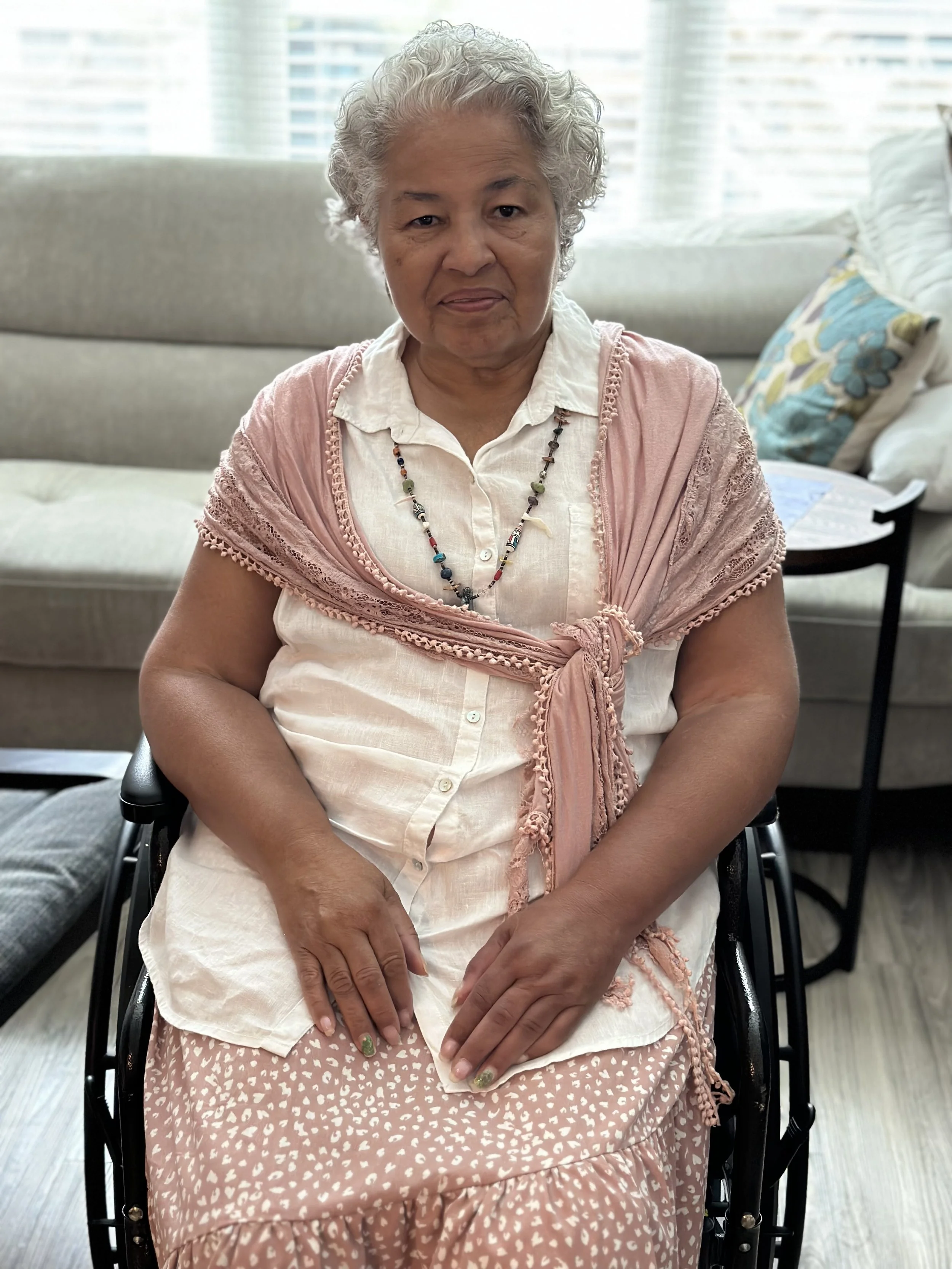 An elderly woman with curly gray hair sitting in a wheelchair indoors, wearing a white blouse, a pink shawl, and a patterned skirt, with a colorful beaded necklace, in a bright living room with a beige couch, decorative pillows, and a window with bli