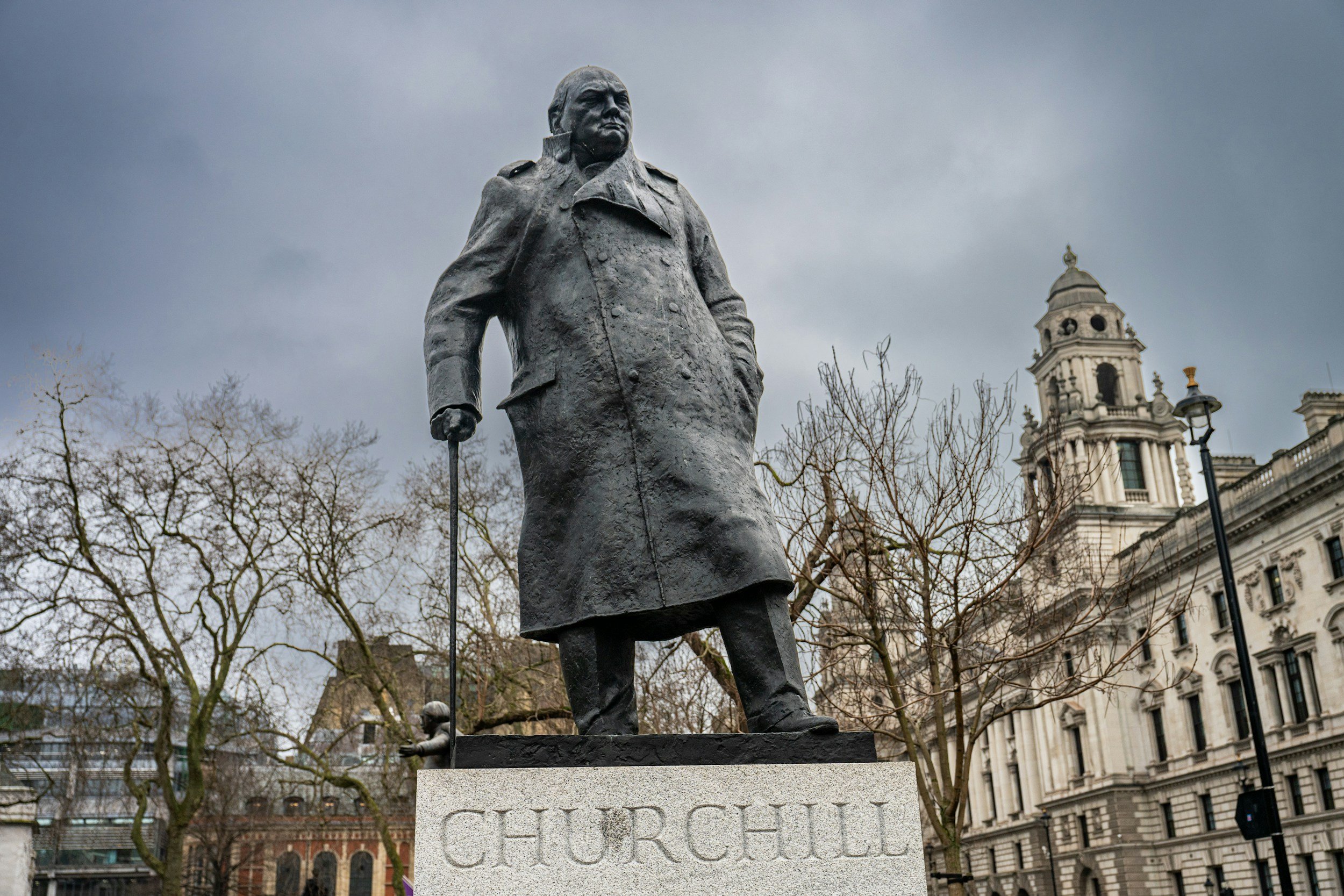 Bronze statue of Winston Churchill on a stone pedestal with his name, in front of a historic building with a dome and baroque architecture, and leafless trees in the background under an overcast sky.