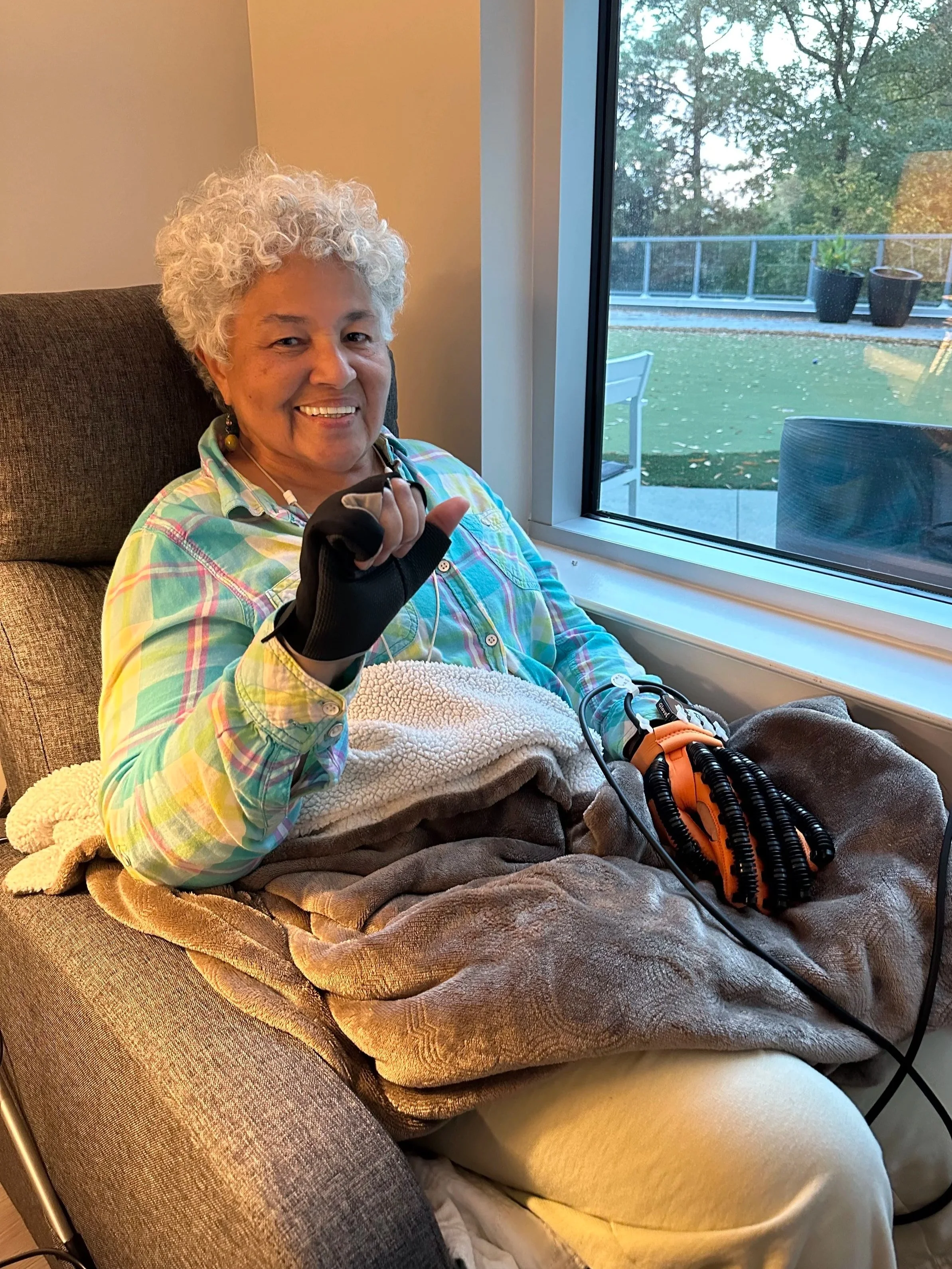 An elderly woman with curly gray hair smiling and making a sign with her hand, sitting in a chair near a large window, with a blanket and medical equipment on her lap.