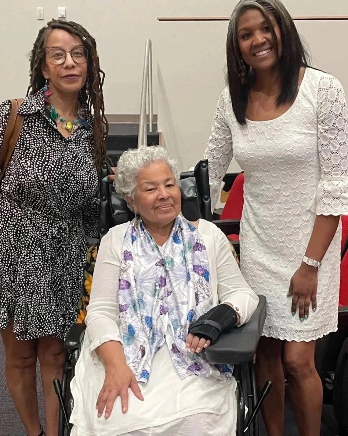 Three women, one sitting in a wheelchair and two standing beside her, smiling at the camera.