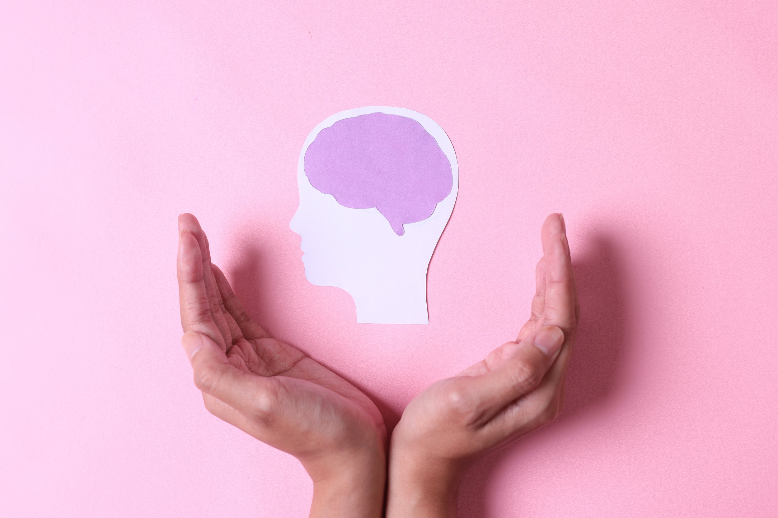 Two hands holding a paper cutout of a human head silhouette with a purple brain inside against a pink background.