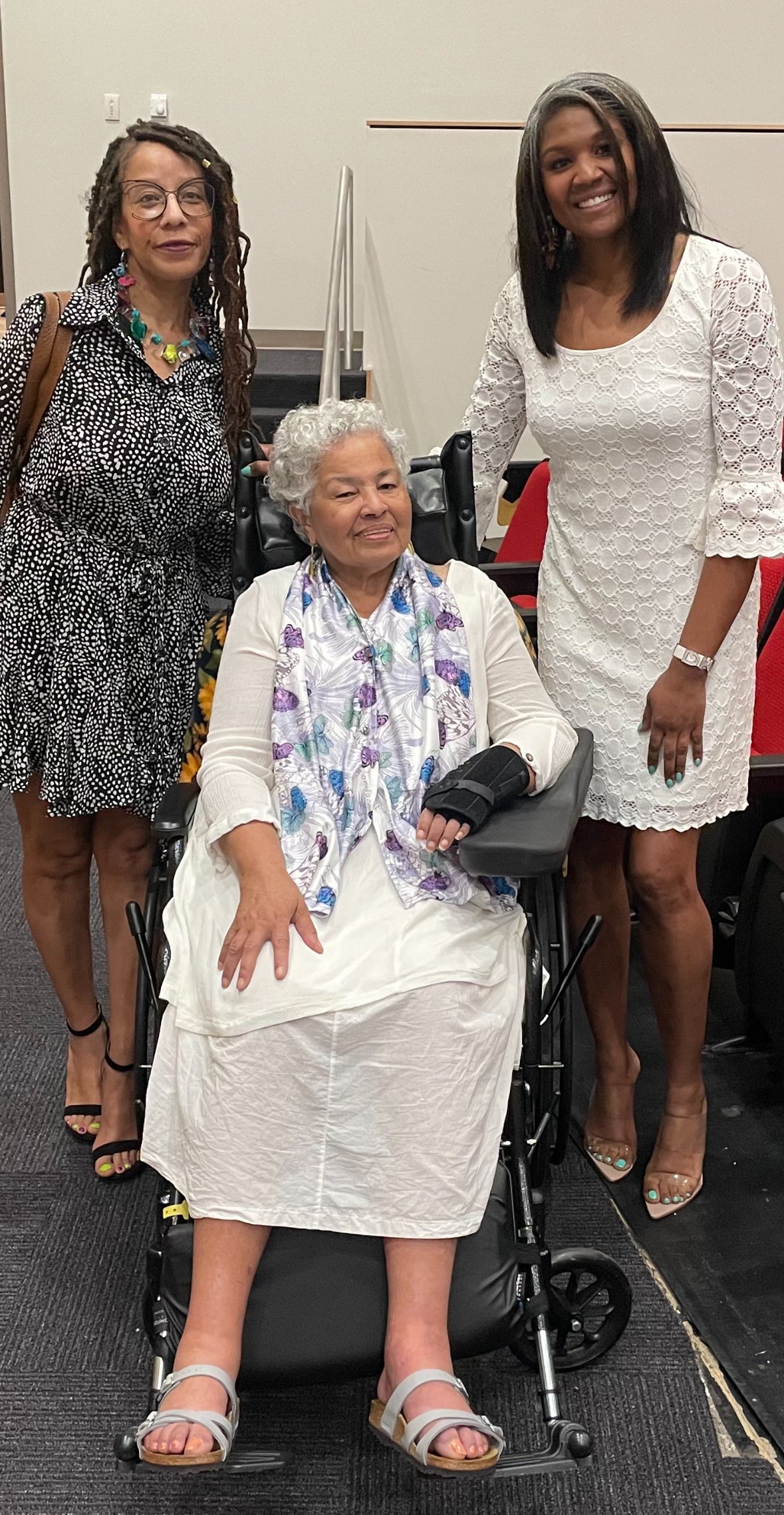 Three women, one sitting in a wheelchair and two standing beside her, smiling at the camera.