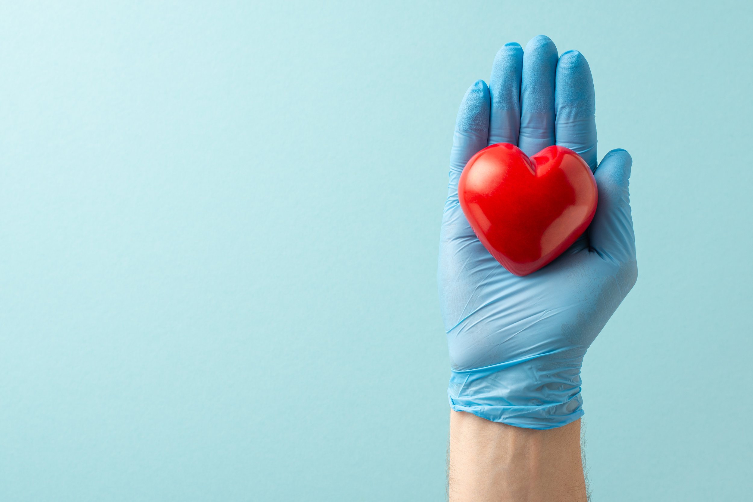 A person's hand wearing a blue glove holding a red heart against a light blue background.