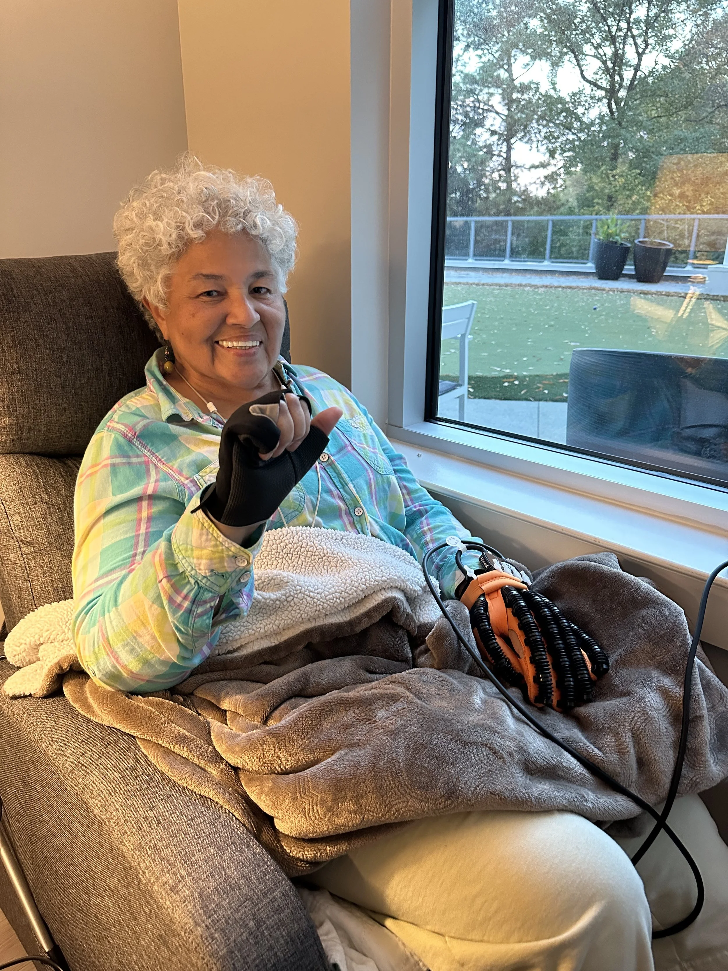 An elderly woman with curly gray hair smiling and making a sign with her hand, sitting in a chair near a large window, with a blanket and medical equipment on her lap.