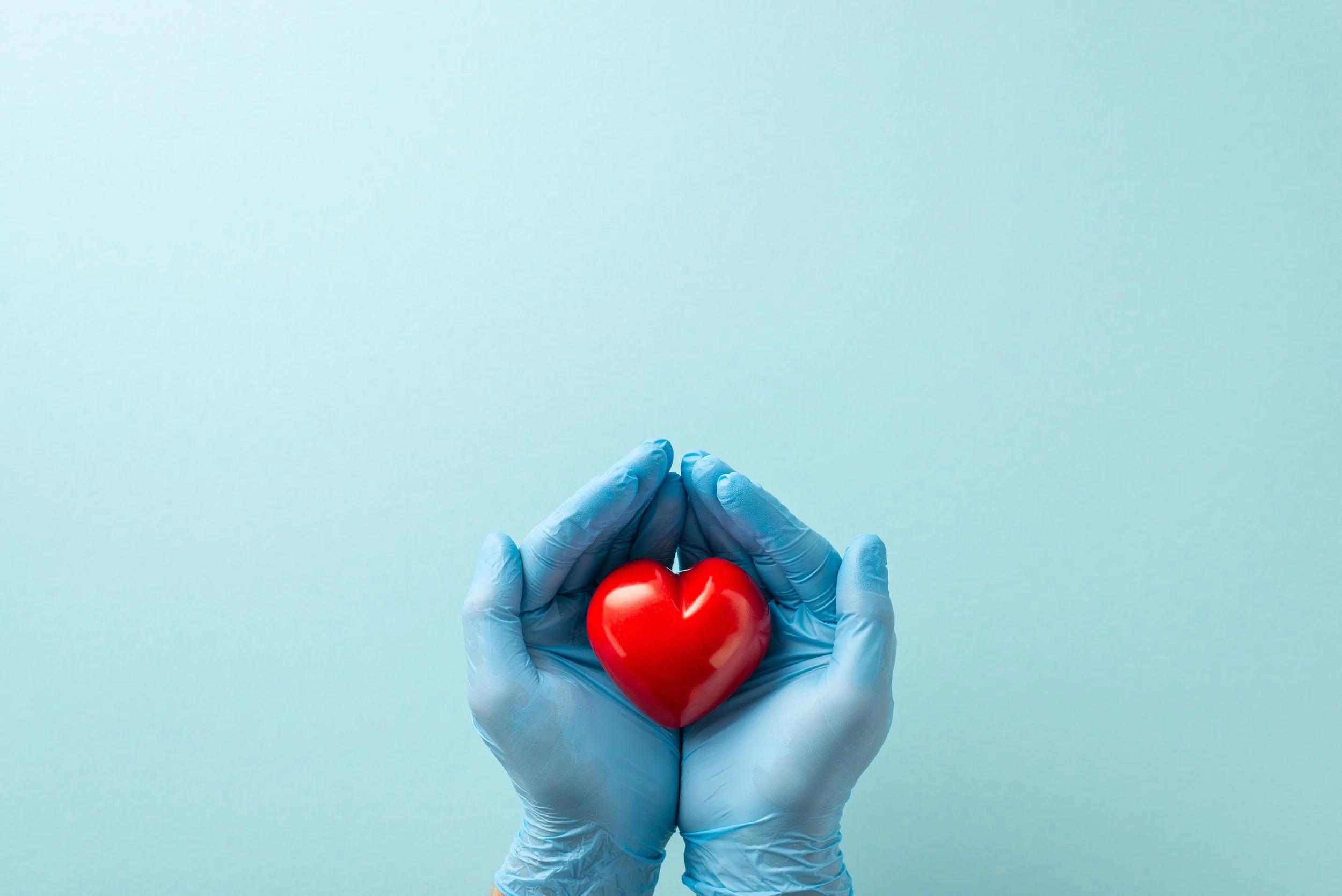 Hands in blue medical gloves holding a red heart-shaped object against a light blue background.
