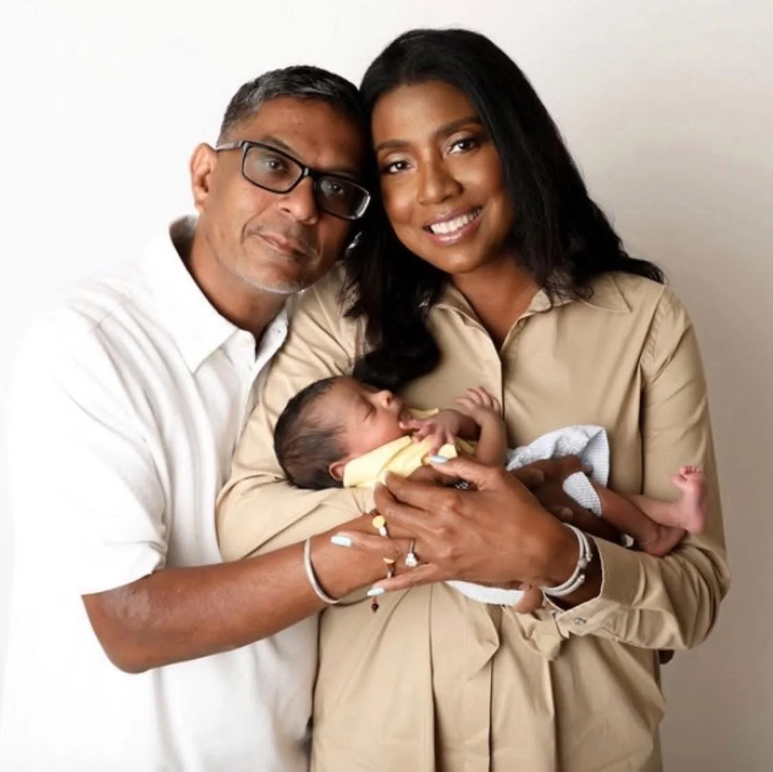 A happy family of three celebrating the birth of their newborn. The father, with glasses, is on the left, and the mother, smiling, is on the right, holding the baby. The baby is sleeping, cradled in the mother's arms.