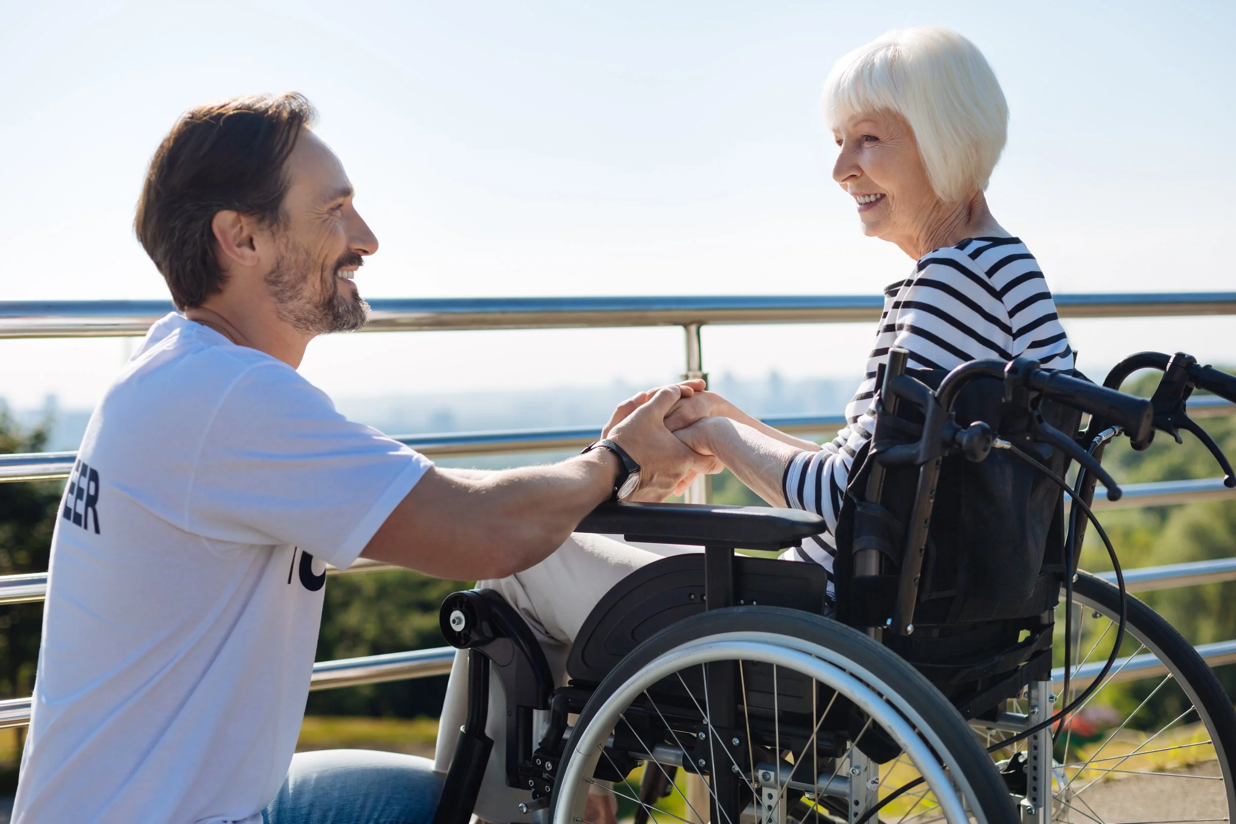 A young man kneeling and holding hands with an elderly woman in a wheelchair, both smiling at each other outdoors on a sunny day with a city skyline in the background.