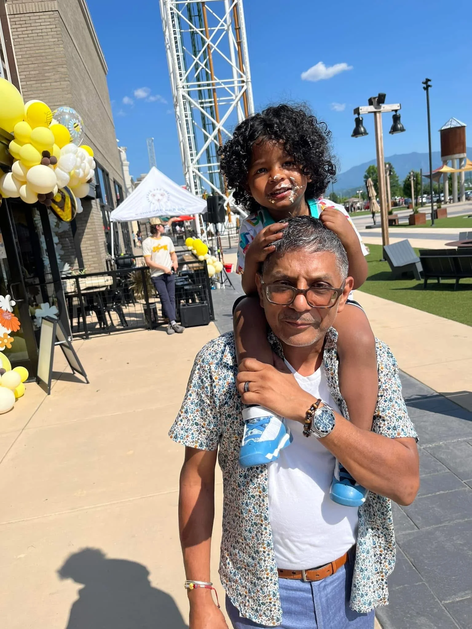 A man carrying a young girl on his shoulders outside on a sunny day, with balloons and a festival or event setup in the background.