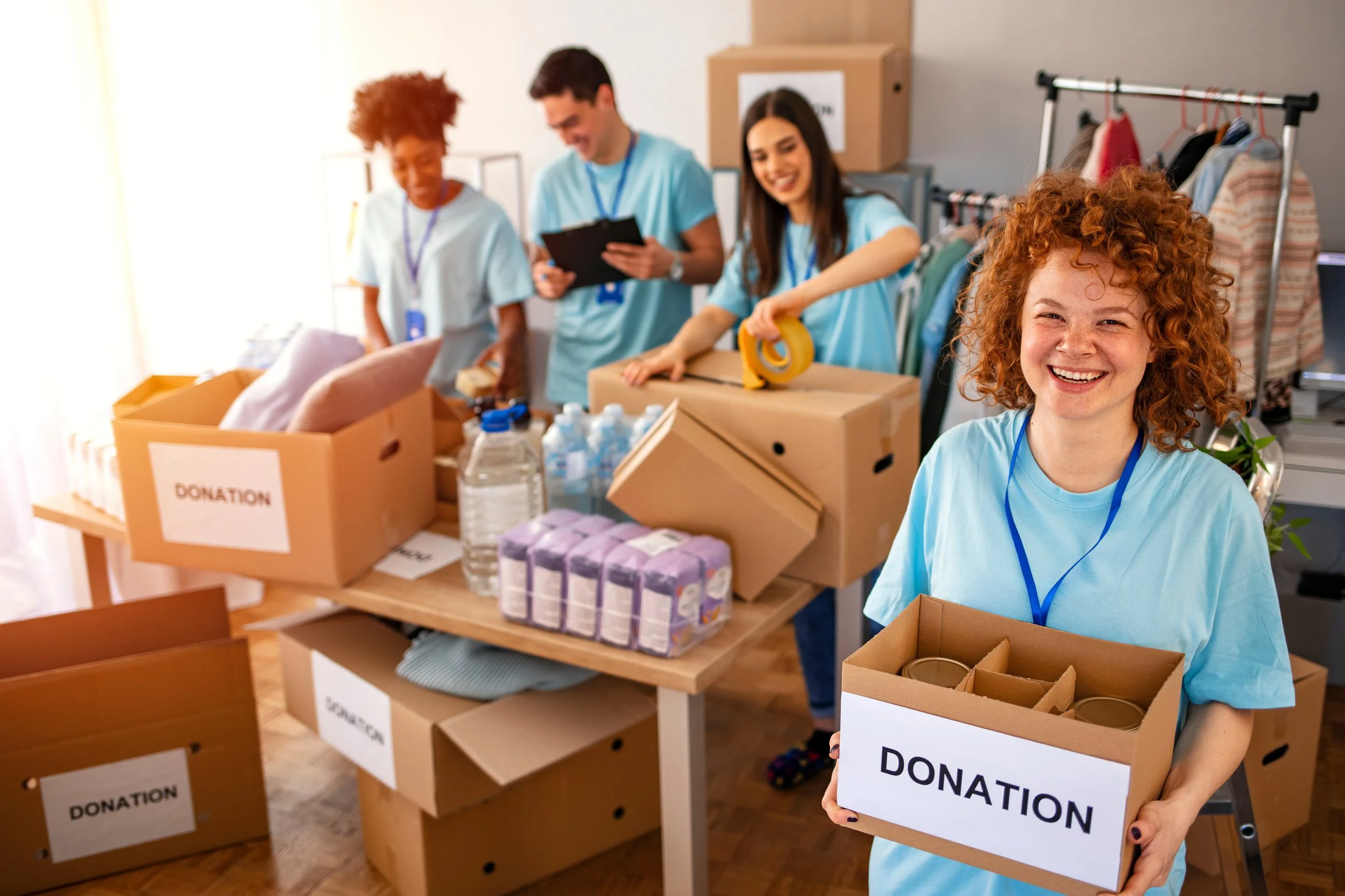 Young volunteers in blue t-shirts packing donation boxes with supplies in a room with coat rack and donation signs.