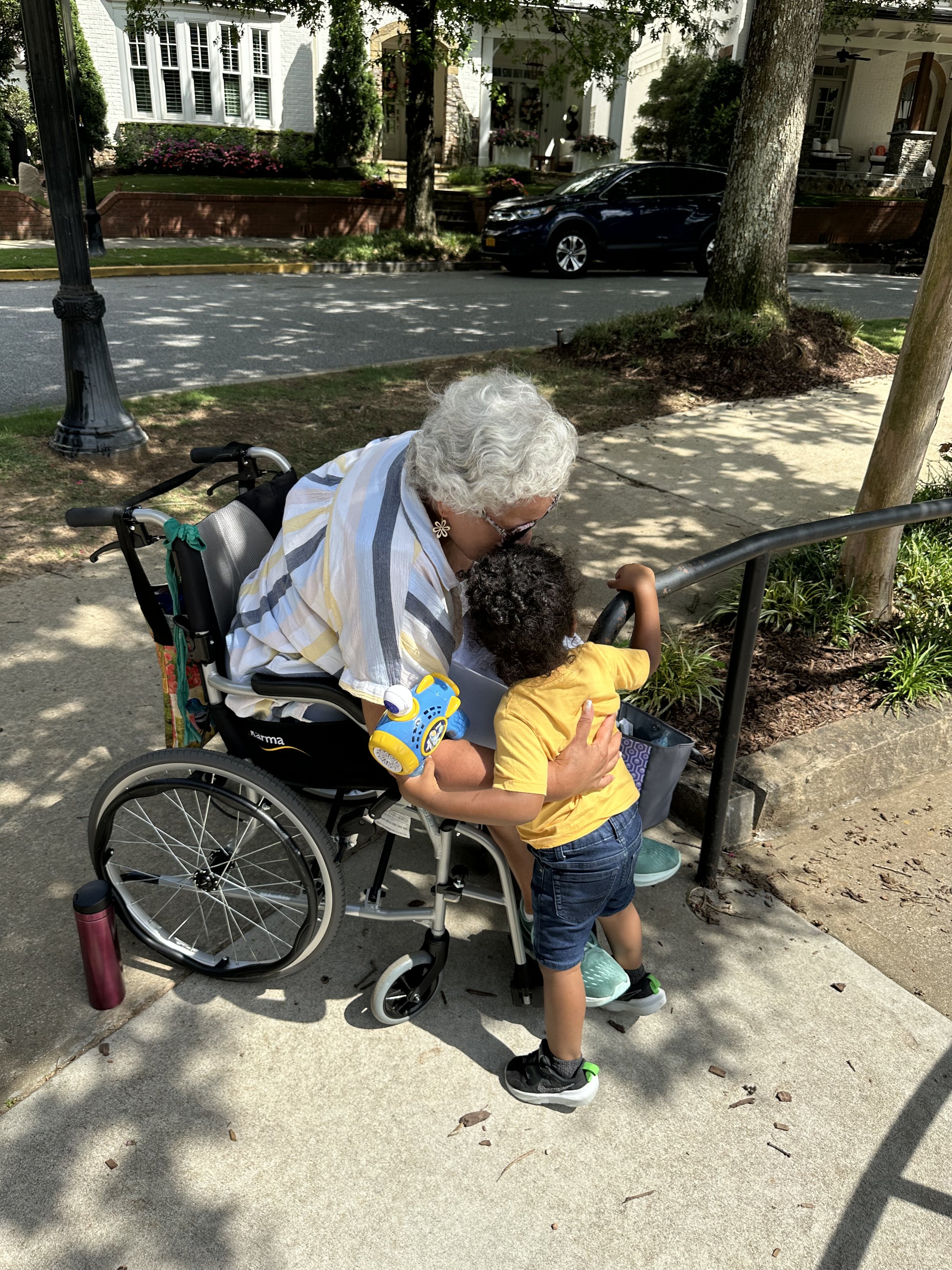 An elderly woman in a wheelchair leaning towards a young child, who is standing on the sidewalk and looking into a small box or container, with trees, grass, and houses in the background.