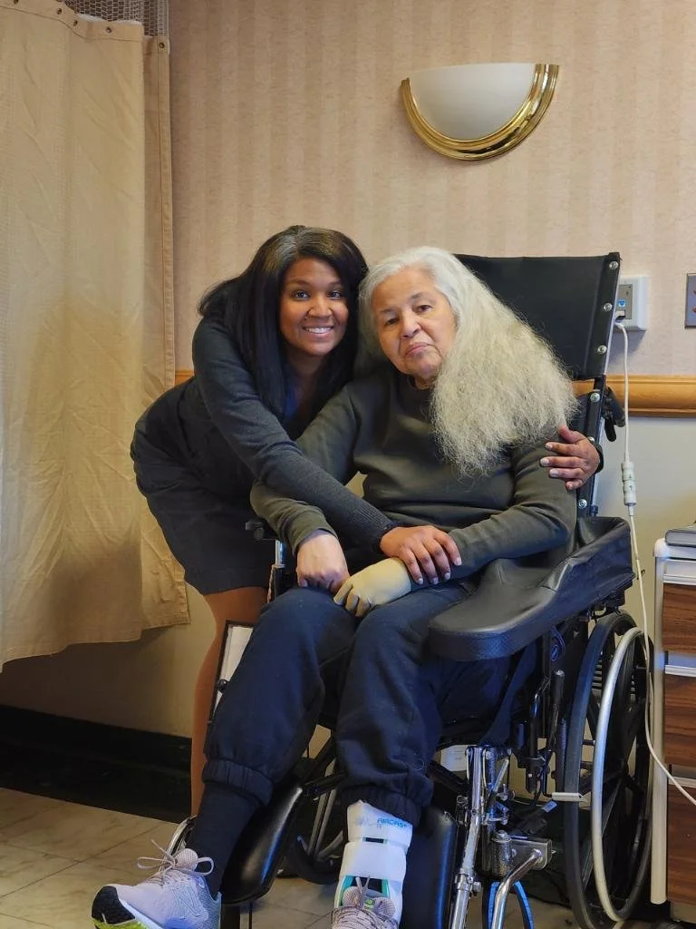 A young woman with dark hair smiling, hugging an older woman with white hair sitting in a wheelchair, indoors in a room with beige walls and a mirror.
