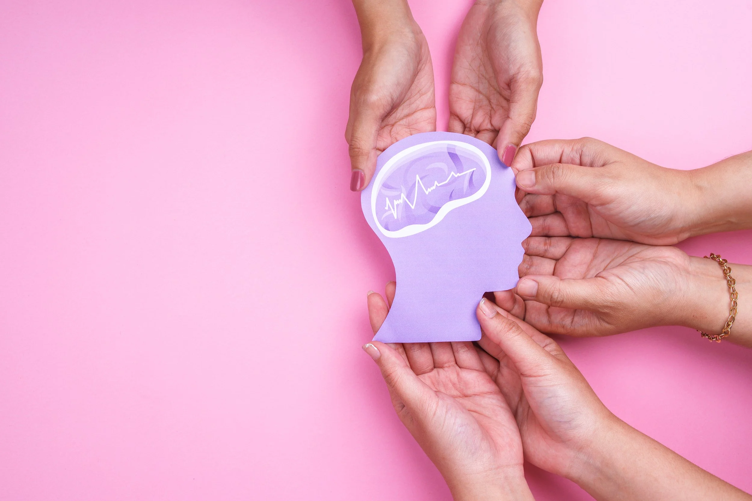 Three hands holding a purple cutout of a human head with a brain illustration and a heartbeat line on a pink background