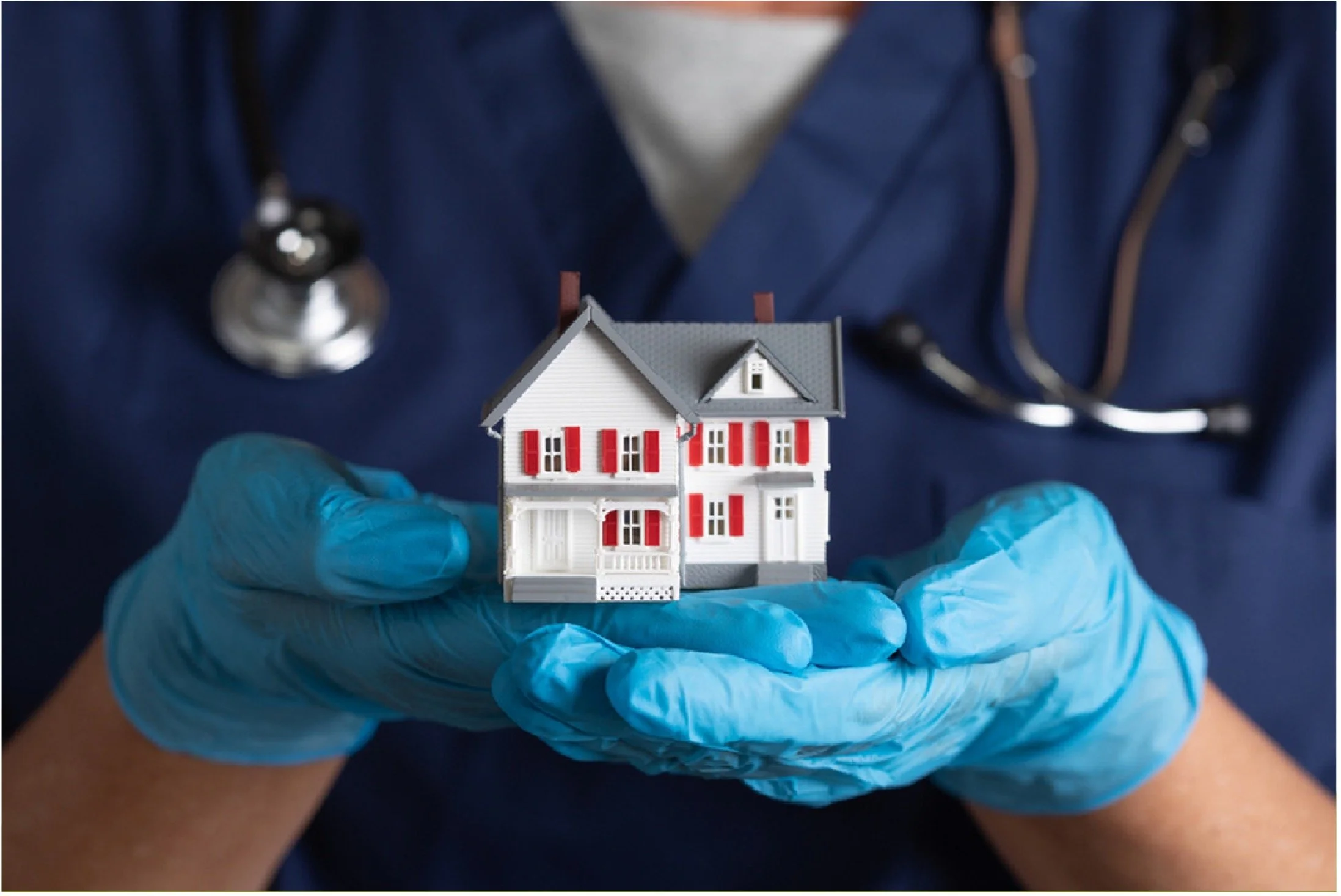 A person wearing blue medical gloves and a navy blue medical jacket holds a small model of a house with red shutters.