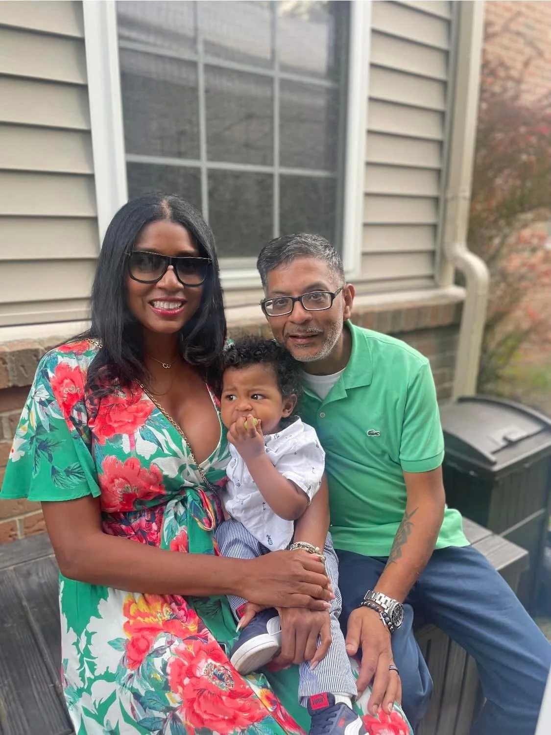 A family of three sitting outside on a wooden deck. The woman is wearing a colorful floral dress, sunglasses, and has long black hair. The man is wearing glasses, a green polo shirt, and has short dark hair. The young boy, sitting on the woman's lap,