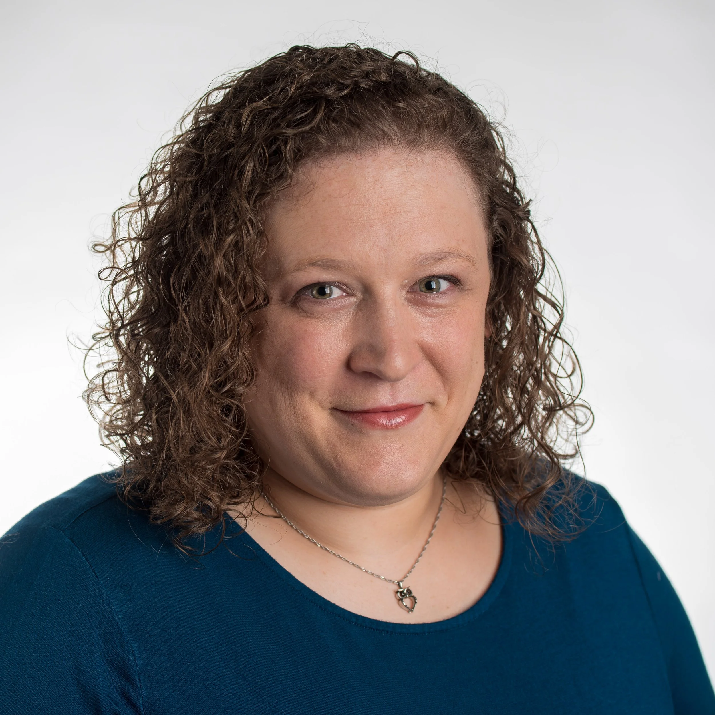 Close-up portrait of a woman with curly brown hair, wearing a blue top and a necklace with a heart-shaped pendant, smiling against a plain white background.