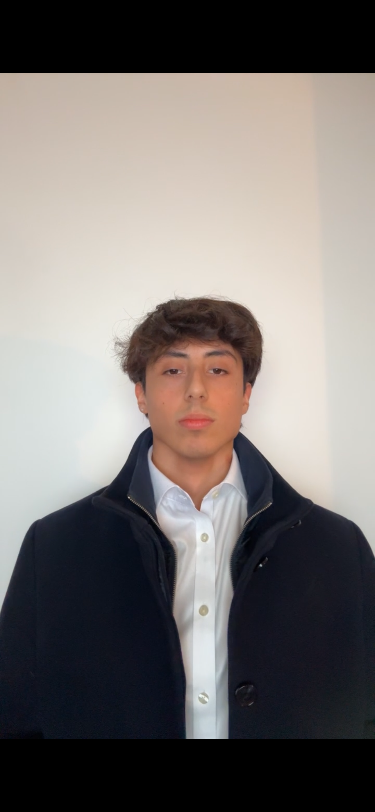 Ung mand med brunt, curly hår iført en sort jakke over en hvid skjorte står foran en hvid væg.