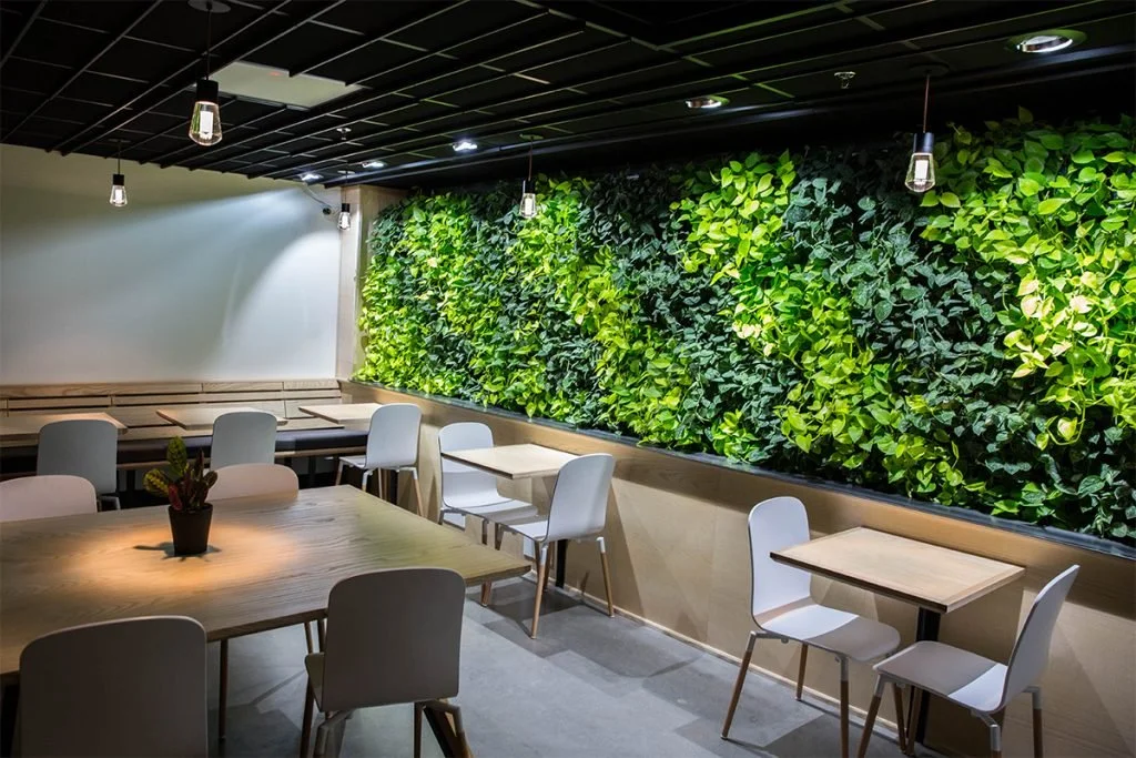 Interior of a cafe or restaurant with wooden tables and white chairs, green wall with foliage, and pendant lighting.