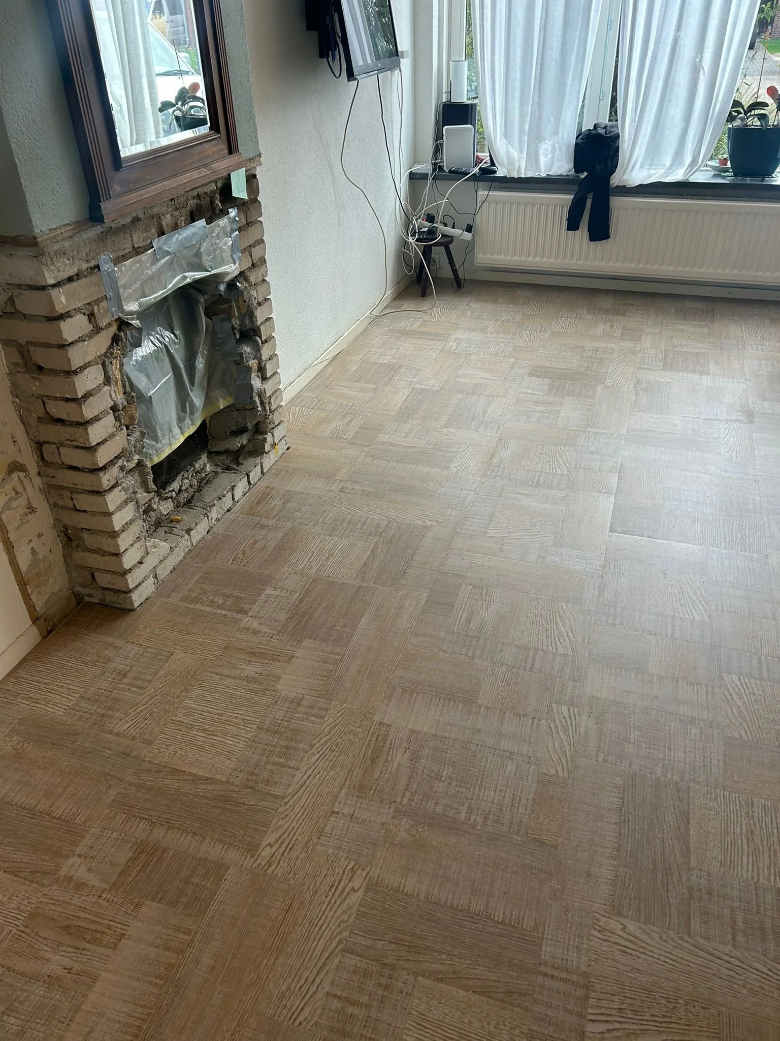 Empty room with a partially installed wooden parquet floor, a brick fireplace without a cover, a wall-mounted TV, and a window with white curtains and potted plants on the windowsill.