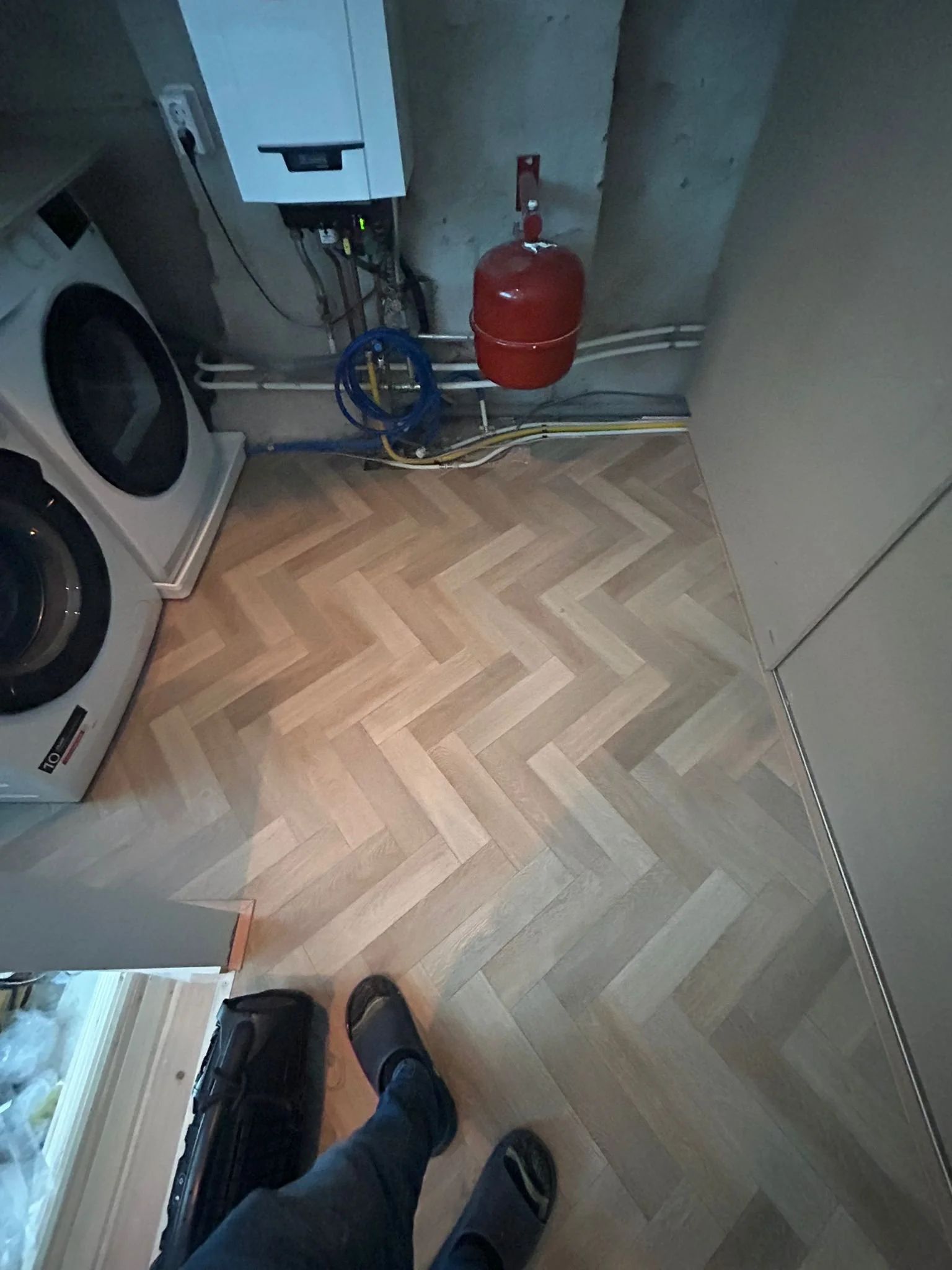 A laundry room with a washing machine, a boiler, a fire extinguisher, and exposed plumbing on a wooden floor.