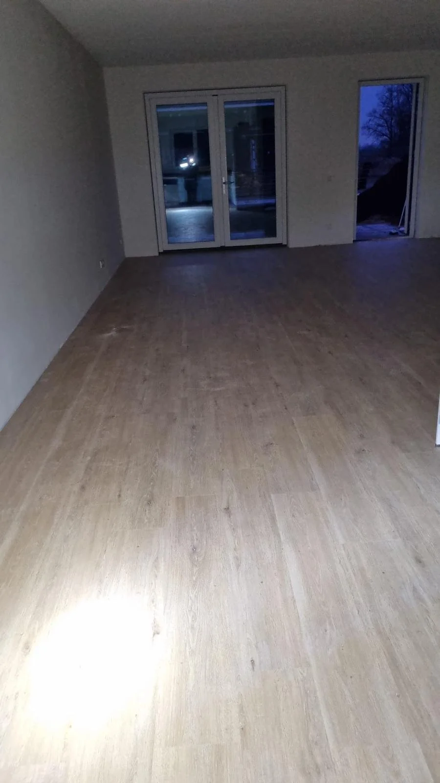 Empty room with wooden flooring, sliding glass doors, and a window opening to the outside, showing snow and dark sky.