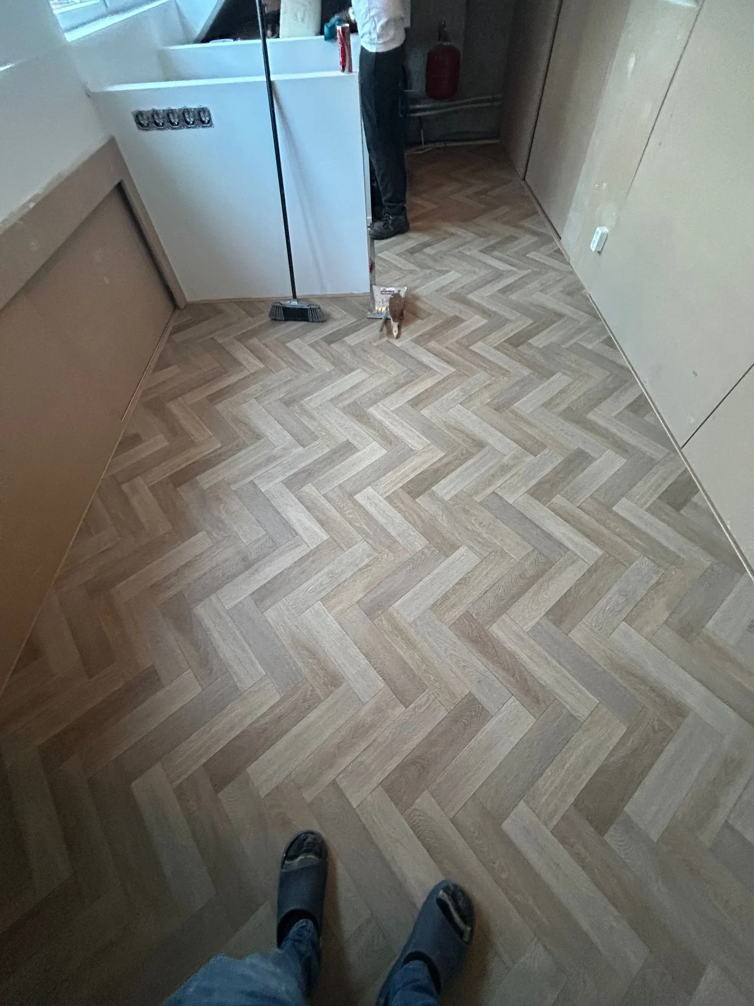 Room with wooden herringbone patterned flooring, a person partially visible standing near a washing machine, cat on the floor, broom leaning against the washing machine, and a person wearing slippers at the bottom of the frame.