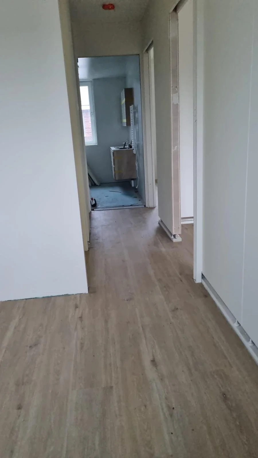 Interior of a home under renovation with wooden flooring, an open doorway leading to a partially finished bathroom, and unfinished door frames.